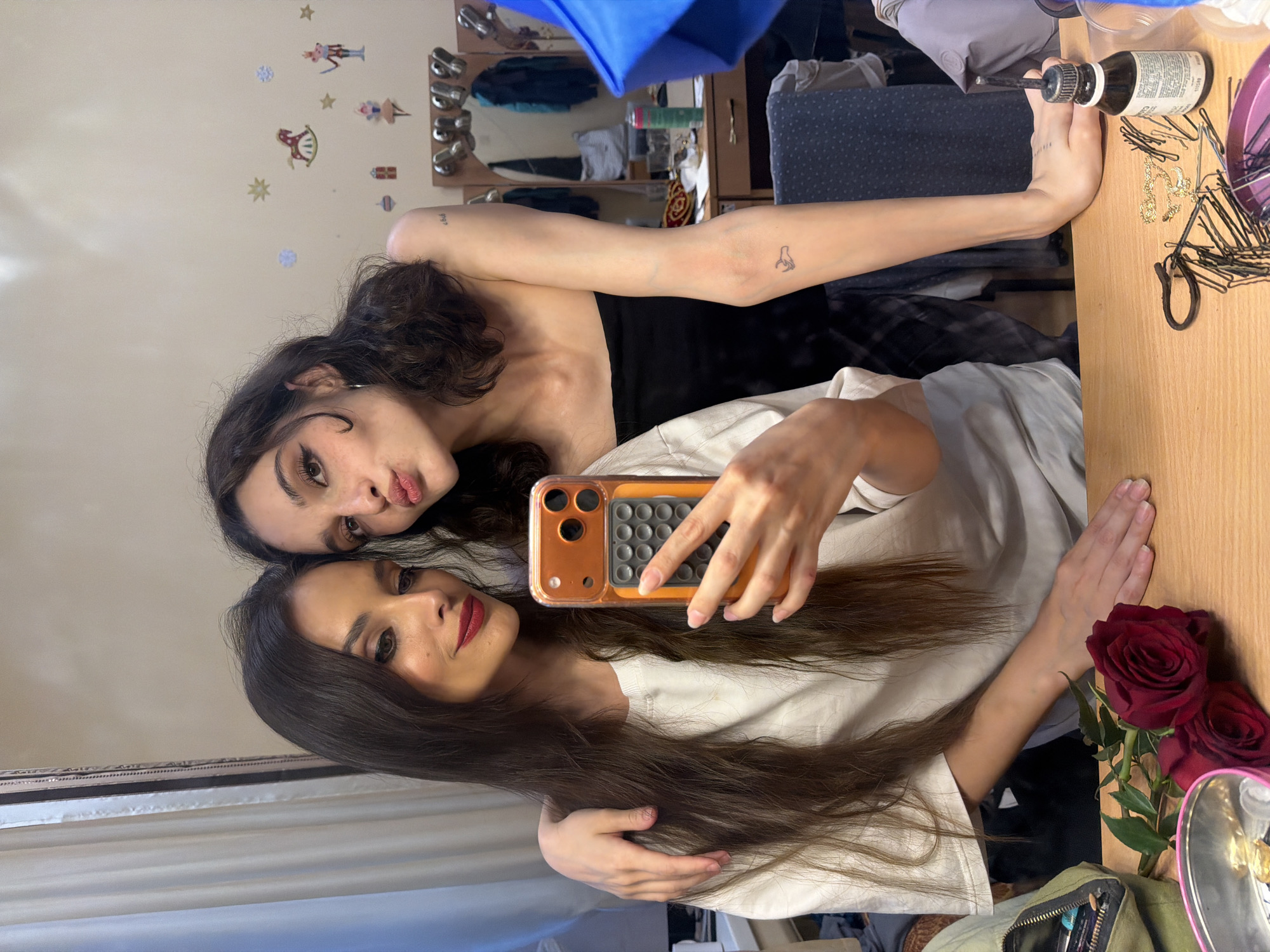 The media file shows two young women taking a mirror selfie in what appears to be a bedroom. One woman is lying down, propped up on her arm, and the other is leaning over her. They are both looking at the camera and smiling. The woman on top has dark, curly hair and is wearing a black top. The woman on the bottom has long, brown hair and is wearing a white t-shirt. She is holding a phone to take the picture.
The background is slightly cluttered, with a mirror visible behind the women reflecting a room with a dresser and various items on it. There are some decorative stickers on the wall, including figures and stars. A bottle with a brush applicator and a pile of bobby pins are visible on a wooden surface, suggesting that the women may be getting ready or doing each other's hair. Two red roses are also placed on the wooden surface, adding a touch of color.
The lighting suggests it could be daytime, possibly indoors. The overall mood seems relaxed and playful, as indicated by the smiles and the casual pose. The location context of Ufa, Russia, is provided, but there are no specific visual cues in the image to confirm this. No legible text is visible in the image.