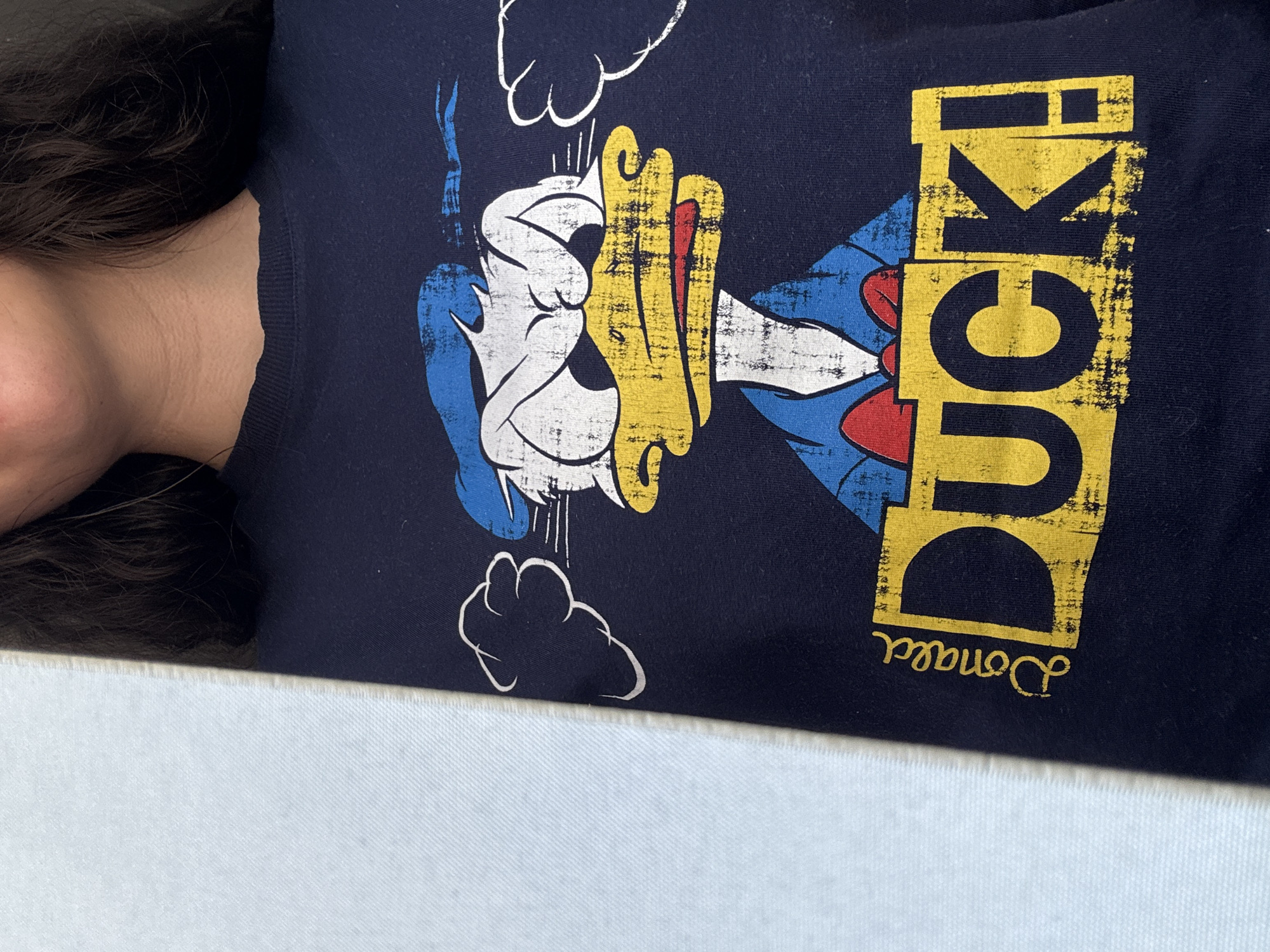 A close-up shot captures a person wearing a dark blue T-shirt featuring a graphic of Donald Duck. The graphic includes the words "Donald Duck" in yellow, arranged vertically. The person's neck and shoulder are visible to the left of the frame, with dark hair partially covering the shoulder. The T-shirt design includes cloud-like shapes near Donald Duck. The background is a plain white surface. The location and city are not discernible from the image.