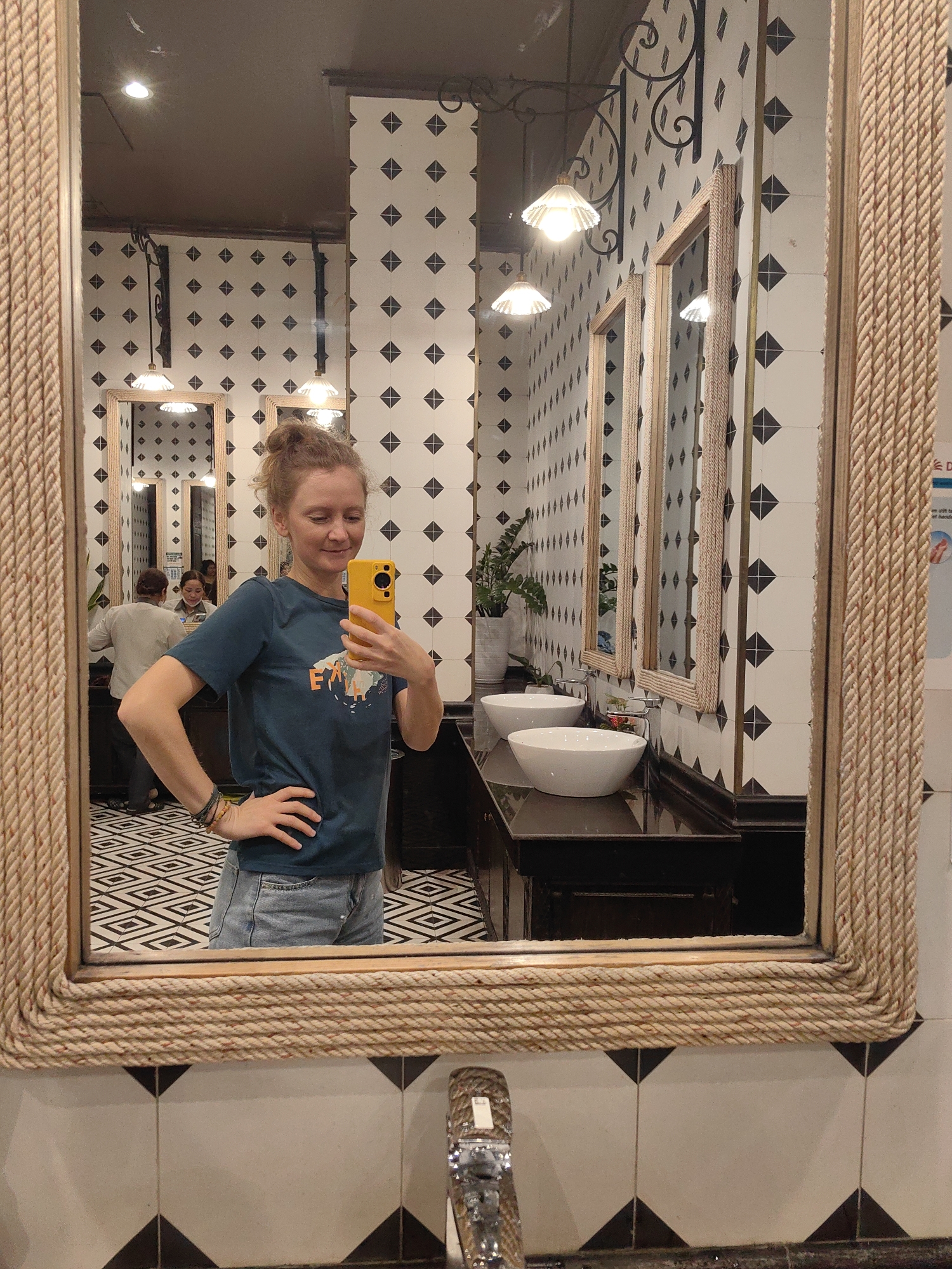 The image depicts a young woman taking a selfie in a mirror. She has curly hair and is wearing a blue t-shirt and denim shorts. The mirror has a rope frame. The background shows a public restroom. The walls are tiled with black and white diamond patterns. There are multiple mirrors above a black countertop with white bowl sinks. Light fixtures hang from the ceiling. Other people are visible in the reflection of the mirror. The image was taken in Bai Chay Ward, Vietnam.