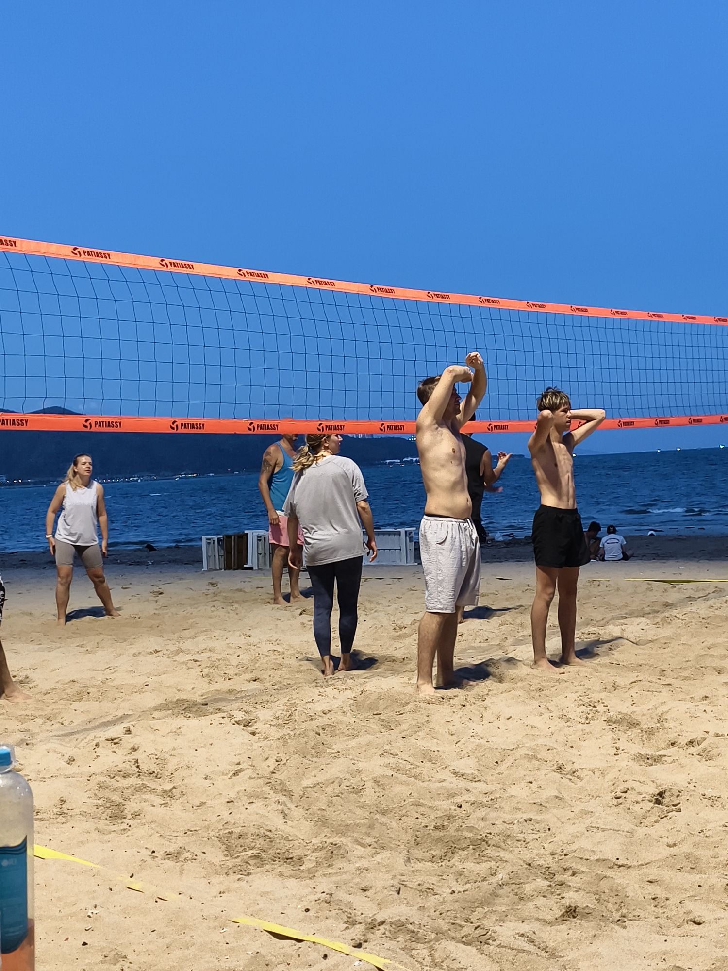 The image shows a beach volleyball game. The setting is a beach in the city of Unknown, Unknown, as implied by the beach, sea, and shoreline visible in the background. A volleyball net with orange borders stretches across the center of the image. Several people are playing volleyball, while others are standing on the beach. One man is shirtless and has his hands raised. Another man is standing next to him, also shirtless with hands behind his head. Another woman is in a white tank top. The sand is a light tan color. In the background, there is the ocean and some land visible in the distance.