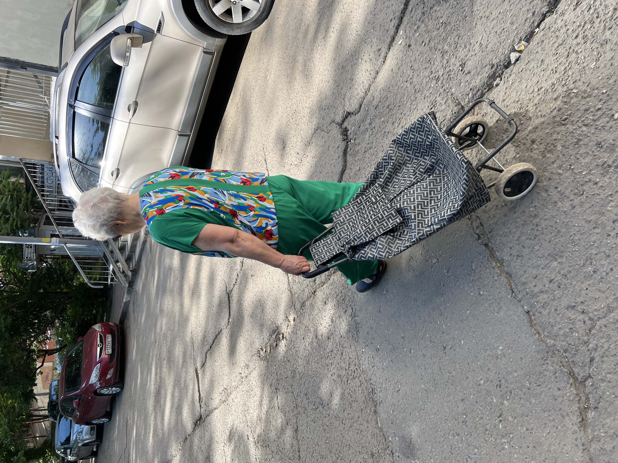 An elderly woman, seen from behind, walks on cracked asphalt pulling a two-wheeled shopping cart in Krasnodar, Russia. She has white-grey hair and wears a vibrant green short-sleeved top with an abstract floral or leaf pattern on the back in shades of red, blue, green, and white, paired with solid green pants and dark sandals. The shopping cart is black and grey with a repeating geometric pattern.
To the left, three cars are parked parallel to the street: a silver sedan, a red Opel Astra with Russian license plate "P 132 BE 193" (confirming Krasnodar Krai), and a dark vehicle partially obscured by trees. To the right, a beige SUV (likely a Chevrolet Captiva) is parked beside a building with metal railings and steps. A sign on the building, partially visible, reads "МАСТЕРСКАЯ" (Workshop) and "НОТНАЯ СТУДИЯ" (Music Studio). The street is asphalt, exhibiting numerous cracks and minor debris. Sunlight from the left casts distinct shadows across the ground and vehicles.