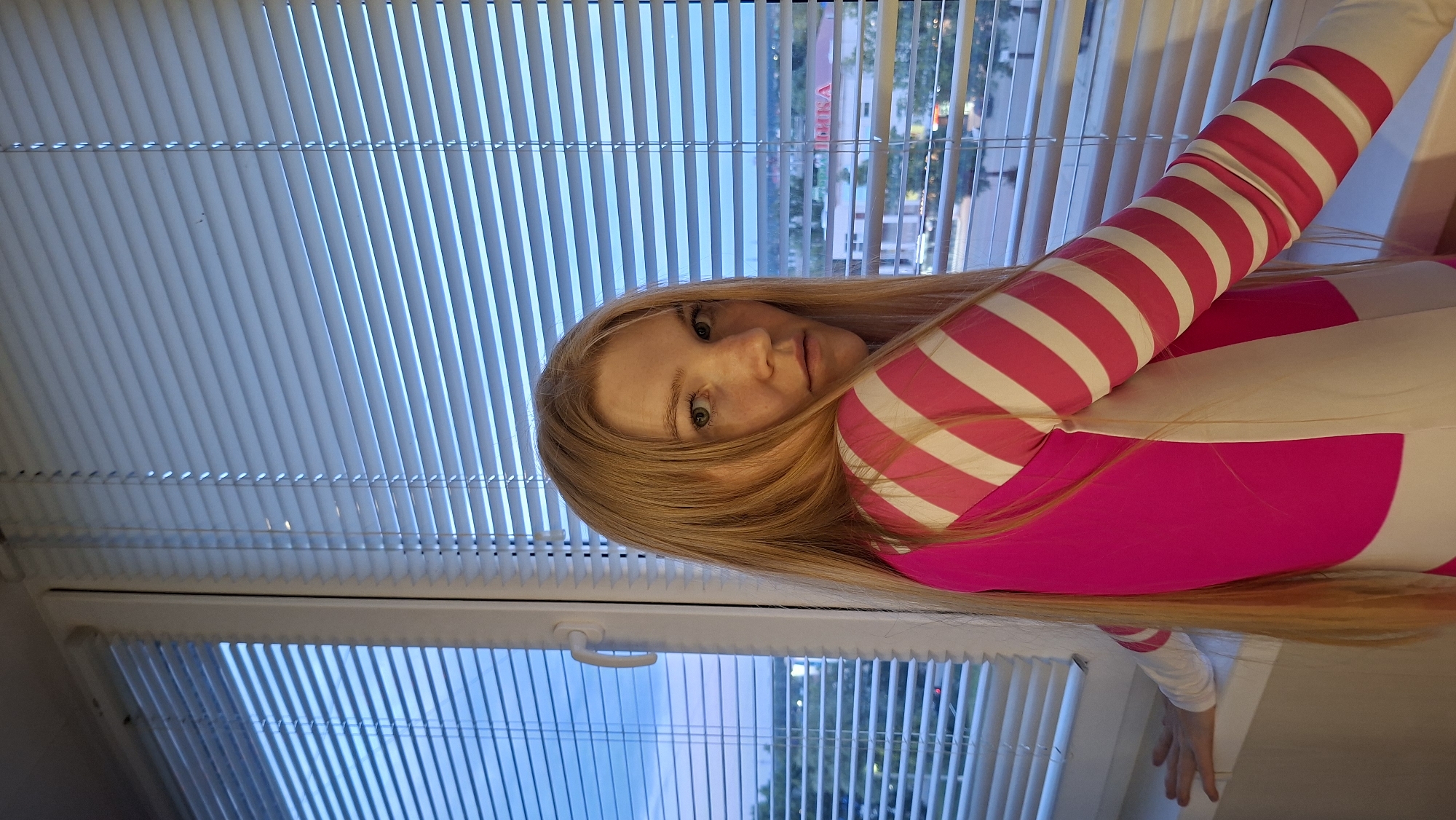 In this media file, a young woman with long blonde hair is captured in a close-up shot, posed sideways. She is wearing a vibrant pink and white striped long-sleeved garment. The background consists of white horizontal blinds, which are partially open, revealing a glimpse of the sky and buildings outside. The lighting suggests it is either dawn or dusk.
The woman's face is turned towards the camera, with her head tilted slightly. Her eyes are open and she has a neutral expression. Freckles are visible on her nose and cheeks.
The setting appears to be indoors, by a window, possibly in a residential apartment. The blinds are a prominent feature of the scene, creating a pattern of light and shadow. The overall impression is a casual and intimate portrait. There is a white window handle visible on the lower part of the frame. The outdoor scene visible through the blinds suggests an urban or suburban environment.
The visible text in the image, partially obscured by the blinds, is "ВЫБИРАЙ" which translates to "CHOOSE" in Russian. This text appears on a building in the background. The location context provided is Chaikovsky, Russia.