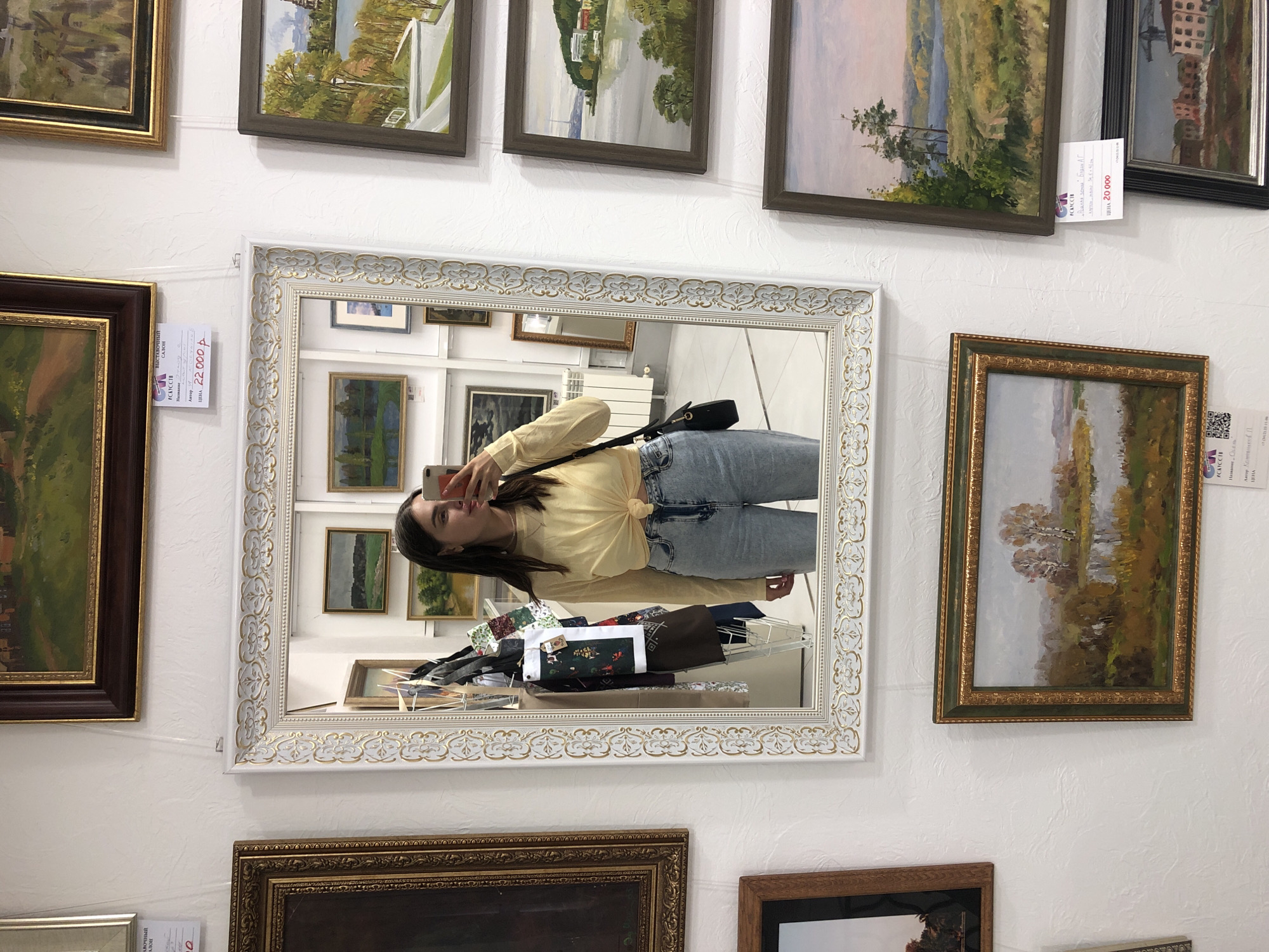 The image shows a woman taking a mirror selfie in an art gallery. She is wearing a yellow long-sleeved shirt tied at the waist and light blue jeans. She is holding a smartphone in her right hand and a black purse is slung over her left shoulder. The mirror is ornate with a white and gold frame. Reflected in the mirror are various framed paintings displayed on the white wall behind her. These paintings appear to be landscapes and still lifes.
The art gallery is located in Izhevsk, Russia. This is indicated by a price tag visible in the upper portion of the image, which reads "ВЫСТАВОЧНЫЙ САЛОН" (Exhibition Salon) and a price in rubles, as well as "ИЖЕВСК" (Izhevsk) written below the artist's name and painting title on another tag. The walls are textured white, and the floor appears to be tiled. The lighting suggests an indoor setting, typical of a gallery. Other artworks are visible on the walls surrounding the mirror, some with price tags and labels in Russian.