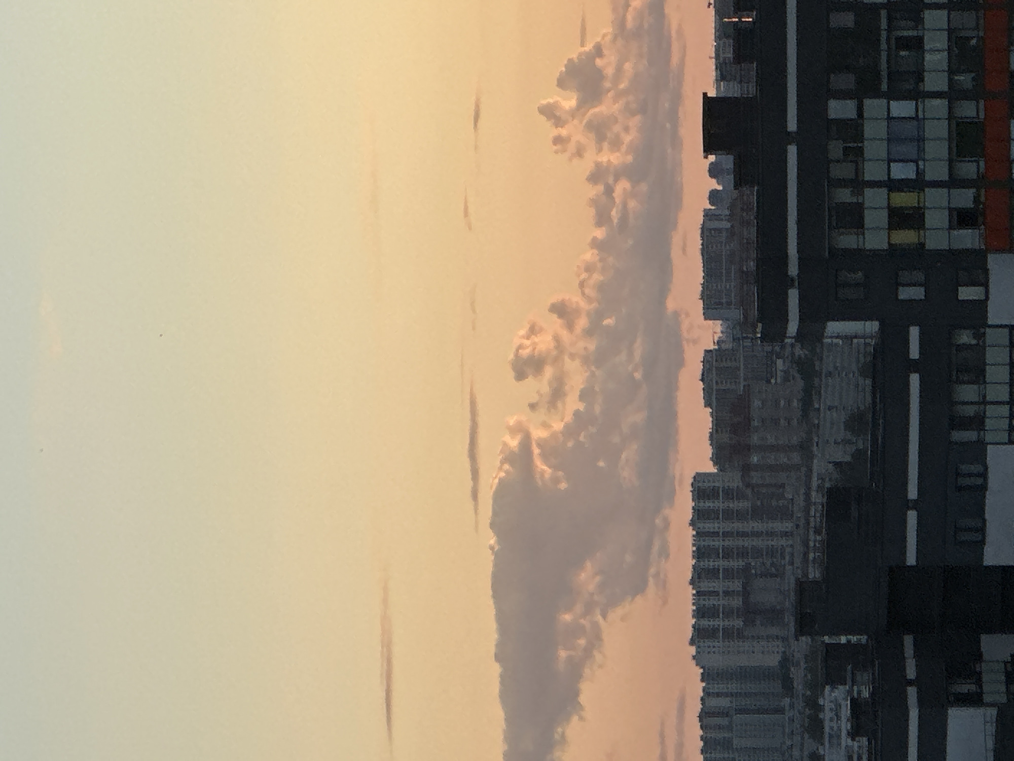 The image depicts a cityscape at sunset, with a prominent sky displaying hues of orange and yellow. A large, textured cloud formation stretches horizontally across the middle of the frame, catching the warm light of the setting sun. Below the clouds, a dense collection of apartment buildings dominates the lower half of the image. These buildings are characterized by their uniform, multi-story structures and appear to be densely packed. The foreground features a dark-colored building with visible windows. In the background, a sprawling urban landscape of apartment blocks extends to the horizon. The overall scene captures an urban environment during twilight. The location is the city of Dnp Ryzhiki, Russia.
