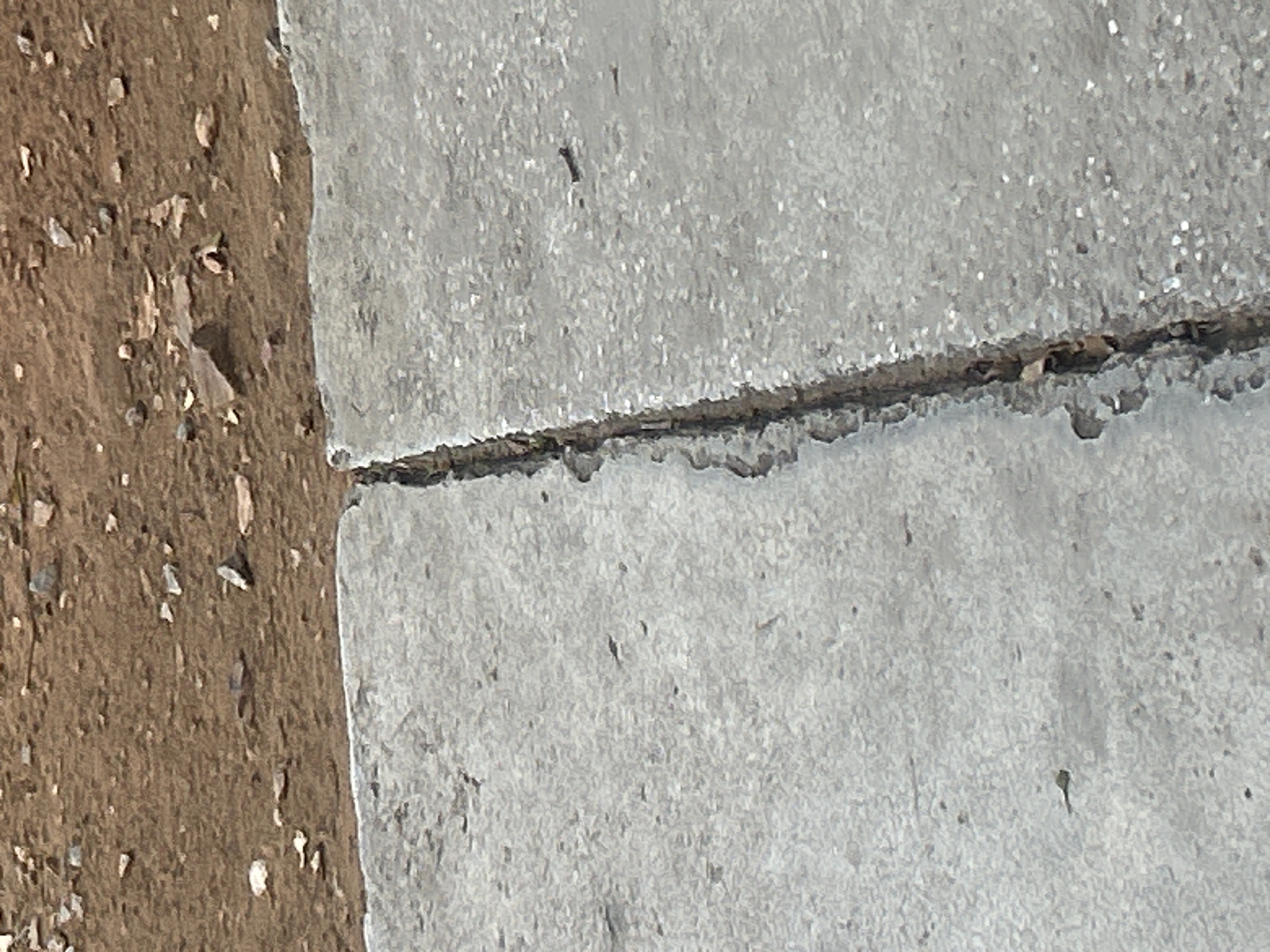 The image shows a close-up view of a crack running through a concrete surface. The concrete is light gray and appears to be weathered, with small pebbles and debris embedded in its surface. The crack is deep and irregular, with darker material, possibly dirt or decaying organic matter, visible within its depths. To the left of the crack, the concrete surface is relatively smooth. To the right, the concrete has a rougher texture with a scattering of small, dark specks, possibly dirt or tiny stones.
Above the concrete, a patch of reddish-brown, dry soil is visible. This soil is coarse and uneven, containing scattered small rocks and pebbles of various sizes and colors, including white, gray, and reddish-brown. Some of these rocks are angular, while others are more rounded. The transition between the concrete and the soil is a sharp, straight line, suggesting a deliberate edge or boundary. There are no people, animals, or vehicles visible in the image. The lighting suggests an outdoor environment under daylight conditions. The specific location in Santa Ana, Venezuela, cannot be determined from the visual information alone.