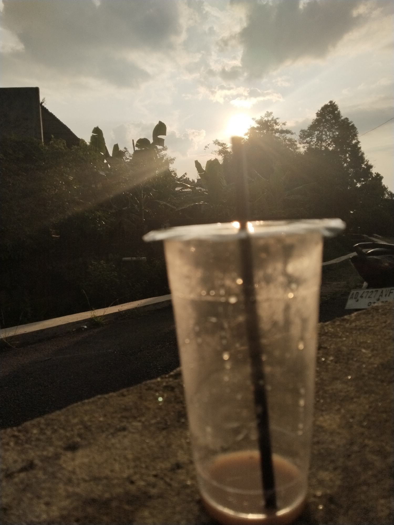A nearly empty plastic cup with a black straw stands in the foreground, positioned on a stone surface. Behind the cup, the scene opens to a view of trees and foliage silhouetted against a bright sky with the sun obscured by clouds. In the middle ground, a concrete curb runs horizontally, and partially visible to the right, is what appears to be a parked motorcycle, with a license plate that reads "AD 4727 AVF". The location seems to be in a residential area, possibly a neighborhood, with dense vegetation.