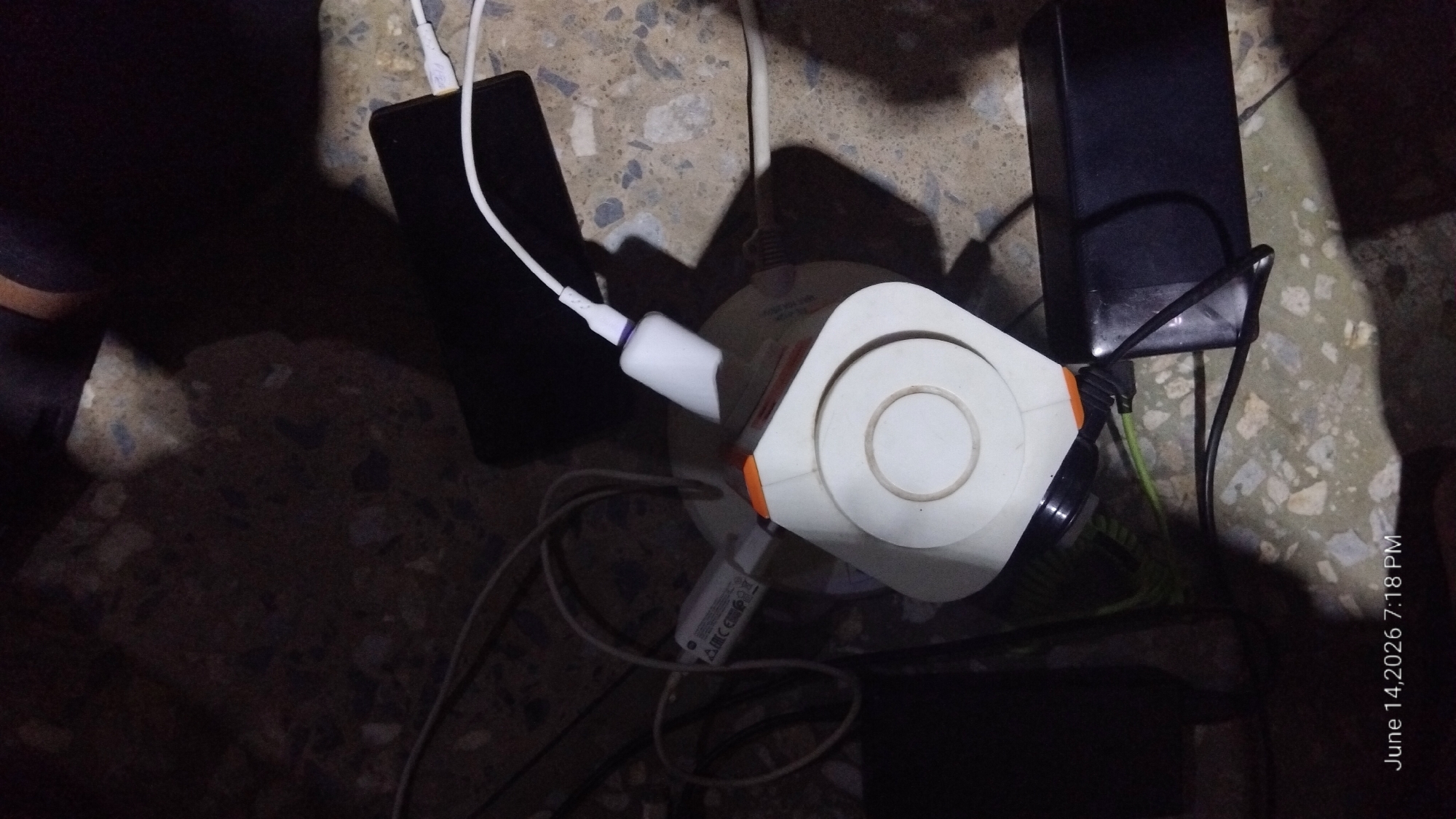 In Howayi, Nigeria, a cluster of electronic devices is arranged on a rough, speckled surface, possibly a floor or a patio. At the center of the arrangement is a white, hexagonal charging hub with orange accents. A white USB charging cable is plugged into the hub, with its other end connected to a black smartphone that is resting on the surface. To the left of the charging hub, a white power adapter with regulatory markings, including "A CE EMC RO HS", is plugged into the hub. Various charging cables, some white and some black, extend from the devices. A black power brick is situated to the right of the charging hub, with a black cable connected to it. A dark, cylindrical object, possibly a battery pack or another adapter, is partially visible to the left of the charging hub. The lighting is dim, casting shadows from the objects onto the textured ground. The timestamp "June 14, 2026 7:18 PM" is visible in the lower-left corner of the image.