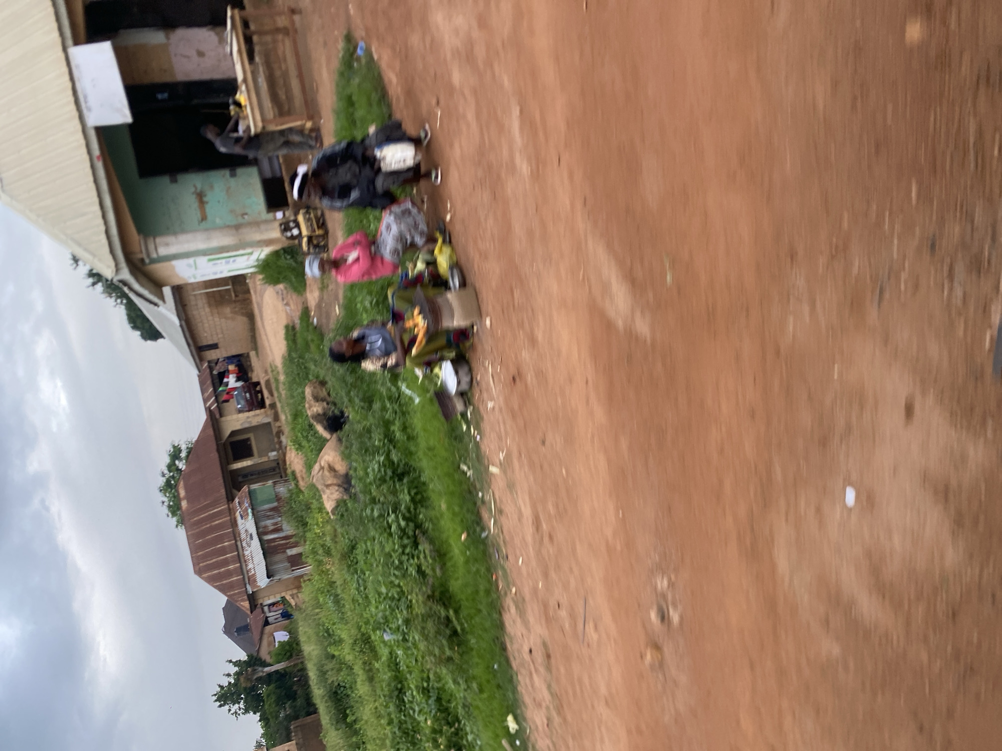 The image depicts a street scene in Jos, Nigeria, under an overcast sky. In the foreground, a dirt road with a reddish-brown hue occupies the majority of the frame, showing some scattered debris. To the left, a patch of lush green grass and vegetation slopes upward, with a few large, brownish rocks or possibly animals interspersed within it.
Several figures are present. Three women are seated on the ground near the grassy area. The woman on the left wears a dark top and a patterned skirt, with corn on the cob arranged in front of her. The middle woman is in a bright pink top and a patterned skirt. The woman on the right is dressed in dark clothing, carrying a white bag, and is seated on what appears to be a stool or crate. In front of them are various items including pots, bags, and what looks like cooking implements, suggesting a street vending setup.
Further back, a man is visible behind a wooden table in front of a building with a light-colored, possibly metal, roof and a green-painted facade. The building appears to be a shop or stall. Other structures, including houses with corrugated iron roofs, are visible in the background, along with a red car parked near one of the buildings. The overall impression is of a casual, everyday street scene in a Nigerian city.