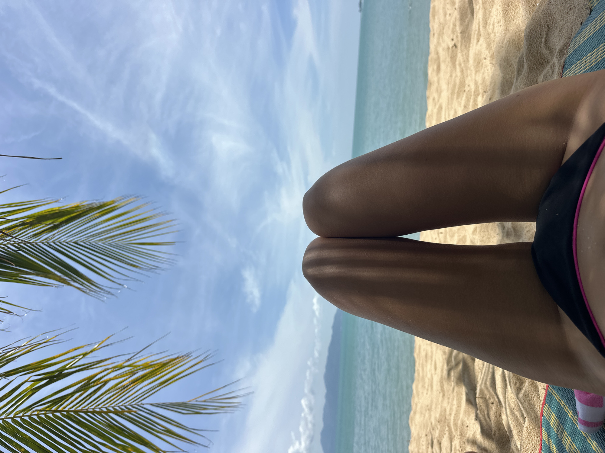 A first-person perspective shot shows a person's legs on a beach in Ko Samui District, Thailand. The person is lying on a colorful mat with stripes of blue, green, and pink. Their lower body is covered by black bikini bottoms with a pink trim. The sandy beach stretches out towards the turquoise ocean, and in the distance, a faint outline of mountains is visible. Palm fronds hang from the top of the frame, casting shadows on the sand and the person's legs, suggesting a sunny day with some cloud cover. The overall scene evokes a relaxed, tropical vacation atmosphere.