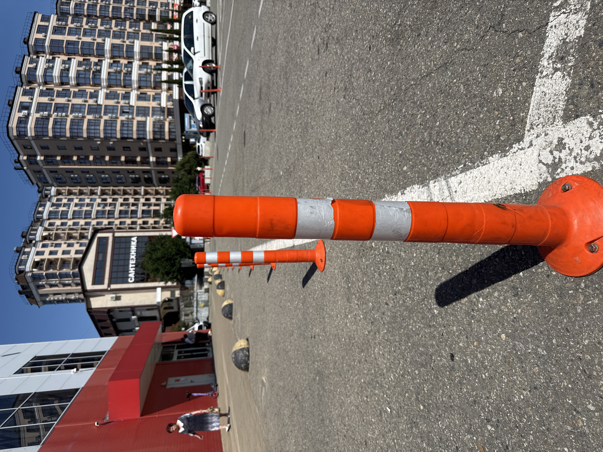 The image, rotated 90 degrees clockwise, captures an outdoor urban scene in Sochi, Russia, on a bright, sunny day with a clear blue sky. The foreground is dominated by a line of orange and white flexible traffic bollards, or road markers, extending across an asphalt parking lot or street. The closest bollard is orange with worn reflective silver/white tape, casting a distinct shadow on the ground, indicating the time of day is likely mid-morning or late afternoon.
In the background, several modern, multi-story buildings with numerous windows and balconies are visible, characteristic of city architecture. Air conditioning units are attached to many of the building facades. A white sedan is parked near one of these buildings, and notably, it has a red wheel clamp or boot on its front wheel. Other vehicles, including another white car and what appears to be a red car, are also present in the background.
A commercial building further back prominently displays the Cyrillic text "САНТЕХНИКА" on its facade, which translates to "Plumbing" or "Sanitary Engineering." Closer to the left of the frame, a large building with red and grey sections is partly visible, and a woman in a dark dress is seen walking, with a purple scooter nearby. Along the asphalt, beyond the main line of orange bollards, there are also shorter, black and yellow spherical bollards. White painted lines on the asphalt demarcate parking areas or road edges.