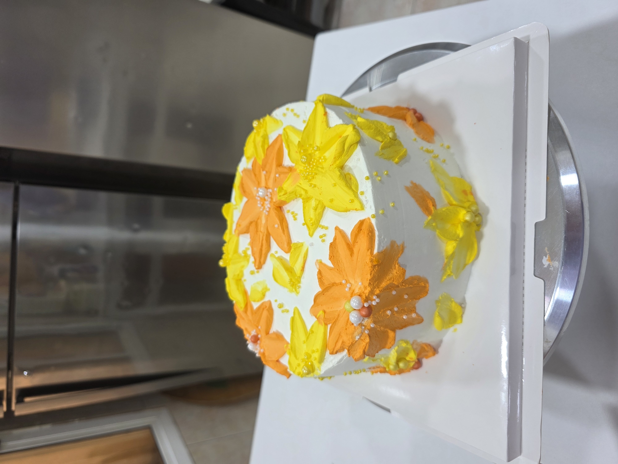 A round cake is positioned on a white cake board, which is then placed on a silver cake stand. The cake is frosted with white buttercream and decorated with an assortment of piped flowers in vibrant shades of orange and yellow. Some of the flowers are adorned with small white and orange edible pearls, and there are scattered yellow sugar pearls across the cake's surface. The background is a blurry indoor setting with a stainless steel appliance, possibly a refrigerator, to the left and a lighter colored wall to the right. The lighting appears to be artificial, casting a soft glow on the cake. There is no indication of the specific city of Dibba Province, Oman.