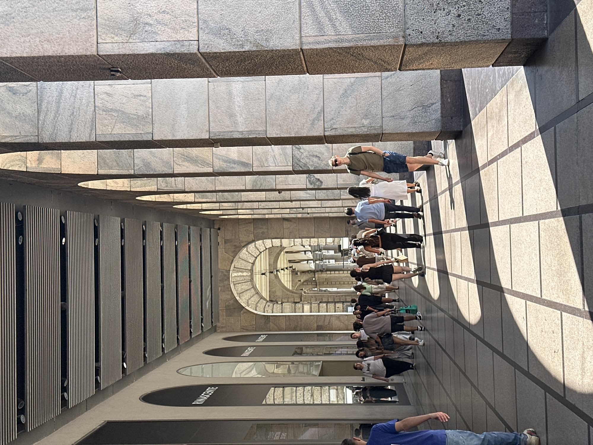 The image depicts a bustling outdoor portico in Milan, Italy, likely within a high-end shopping area. The architecture features grand stone columns supporting a vaulted ceiling, with arched openings revealing the continuation of the portico further down. Sunlight casts strong, geometric shadows across the tiled walkway, highlighting the interplay of light and shade. A diverse group of people, dressed in casual summer attire, are seen walking along the portico. Some are engaged in conversation, while others are focused on their path or the shops lining the walkway, one of which displays the "Rinascente" logo. The atmosphere is one of urban activity on a bright, sunny day.