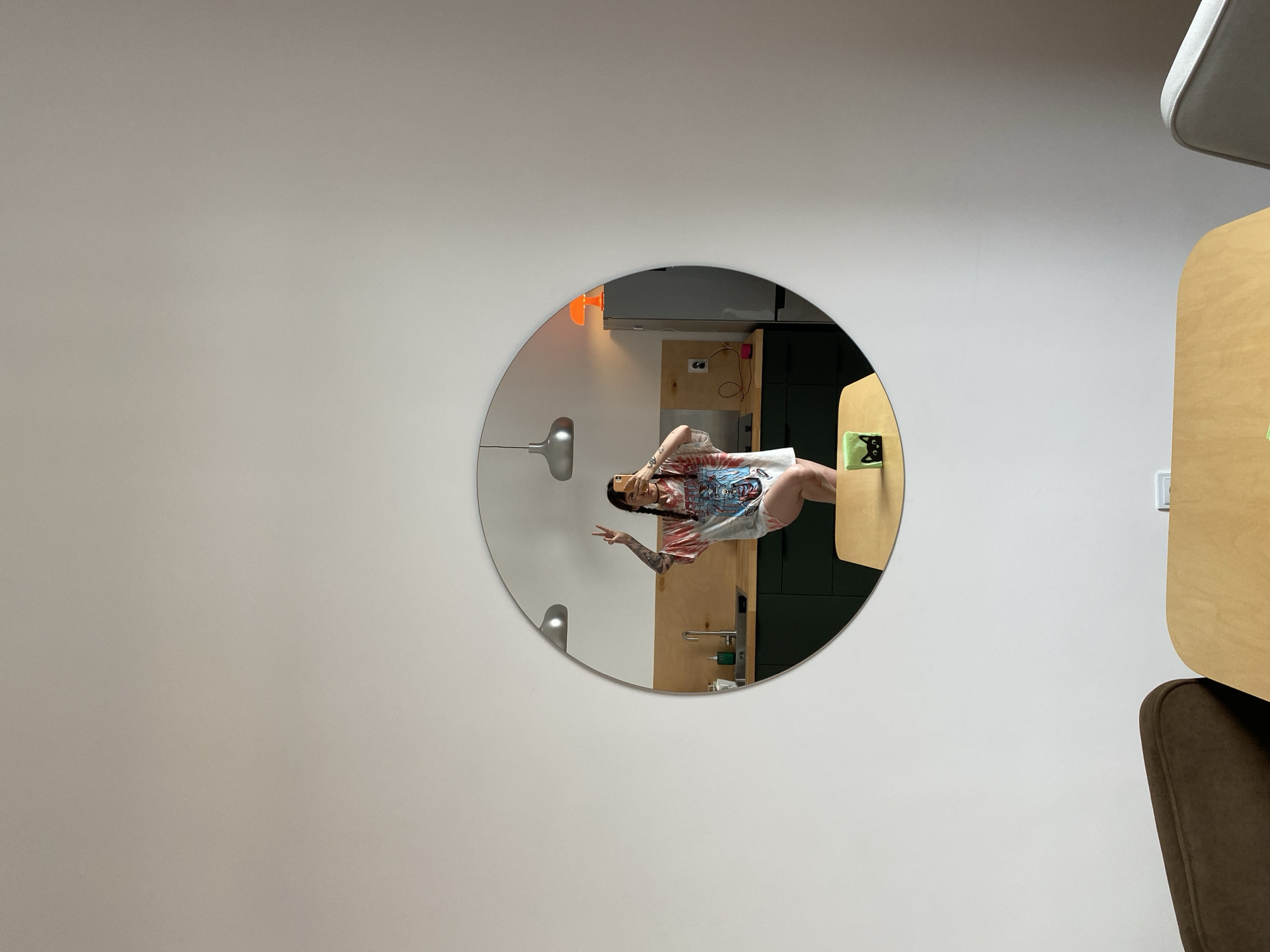 A person is taking a mirror selfie in a modern apartment kitchen. The person is wearing a t-shirt and shorts, and has tattoos on their arms. They are making a peace sign with their left hand and holding a phone in their right hand. The kitchen has dark green cabinets, wooden countertops, and stainless steel appliances. There is an orange lamp on top of the refrigerator. The mirror is round and hangs on a white wall. In the foreground, there is a wooden table with a small black cat figurine on it. This scene is not specifically identifiable to Izhevsk, Russia.