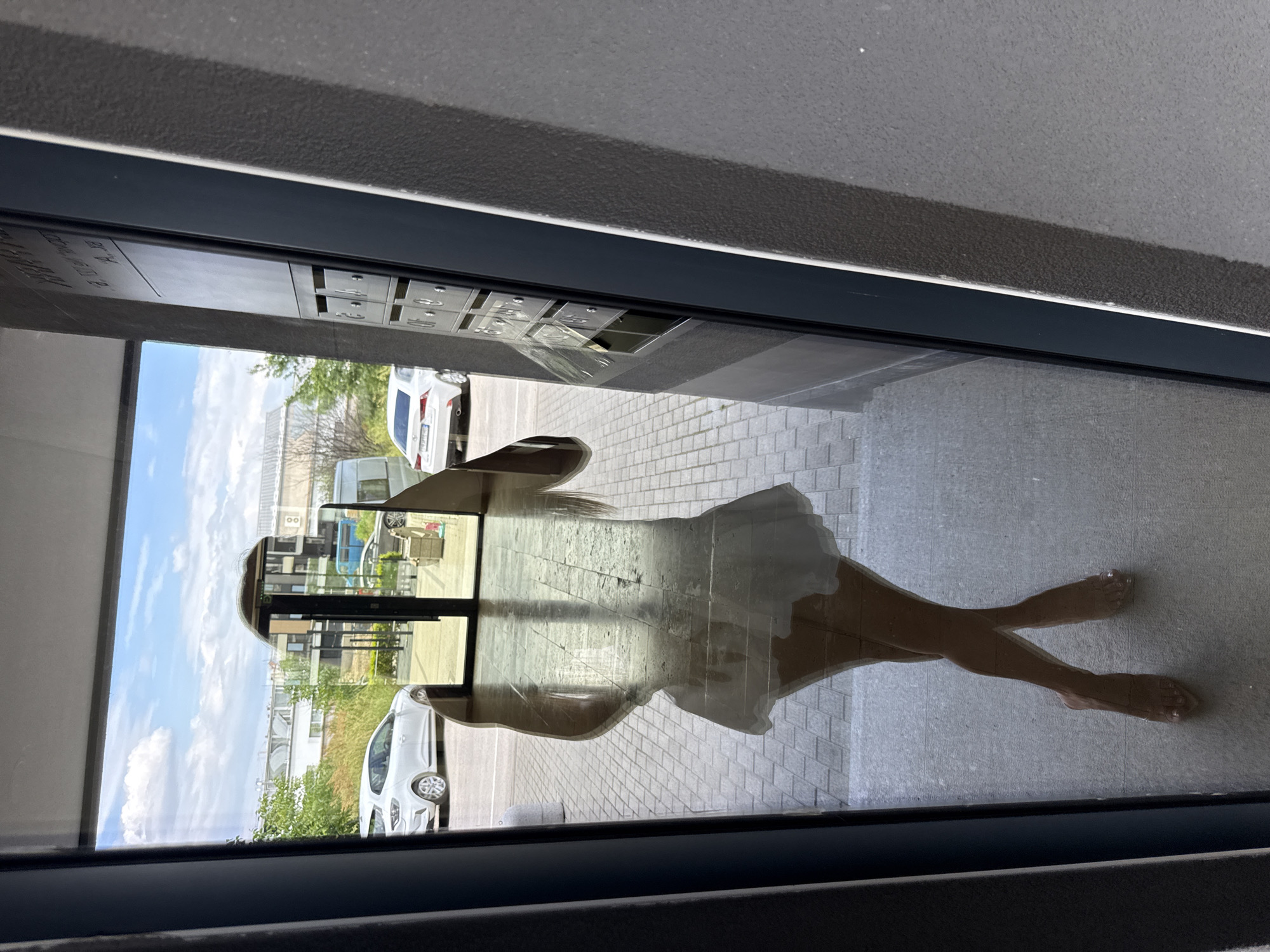 A distorted reflection in a window shows a person in a white dress, with crossed legs, standing in front of the window. The reflection also reveals a cityscape with buildings, trees, cars, and a blue sky with clouds. In the foreground, a concrete structure with mailboxes is visible. The exact location in the city of Unknown, Unknown is not discernible from the image.