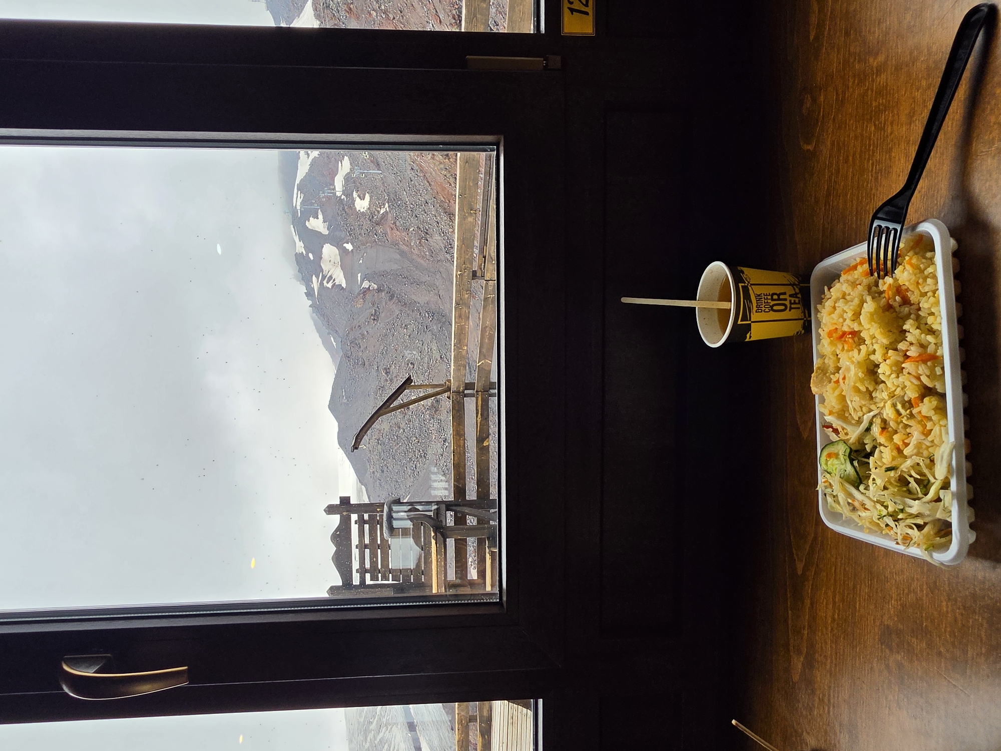The image depicts a meal set on a wooden table, viewed through a window overlooking a mountainous landscape in Terskol, Russia. In the foreground, a white plastic container holds a portion of rice with vegetables and what appears to be a side salad. A black plastic fork rests on the edge of the container. To the right of the food, a yellow paper cup with the text "DRINK COFFEE OR TEA" contains a beverage and a stirring stick.
The window frames a view of a rugged, rocky mountainside, partially covered with snow. A ski lift or cable car system is visible ascending the slope, suggesting a ski resort or a location associated with mountain activities. The sky is overcast and appears to be a pale gray. Outside the window, there is a wooden railing and a rustic wooden chair or bench. The interior of the room is dimly lit, with the table surface reflecting the ambient light. A yellow sign with the number "12" is visible on the right side of the frame, near the window. The overall scene suggests a meal break at a scenic location in the mountains.