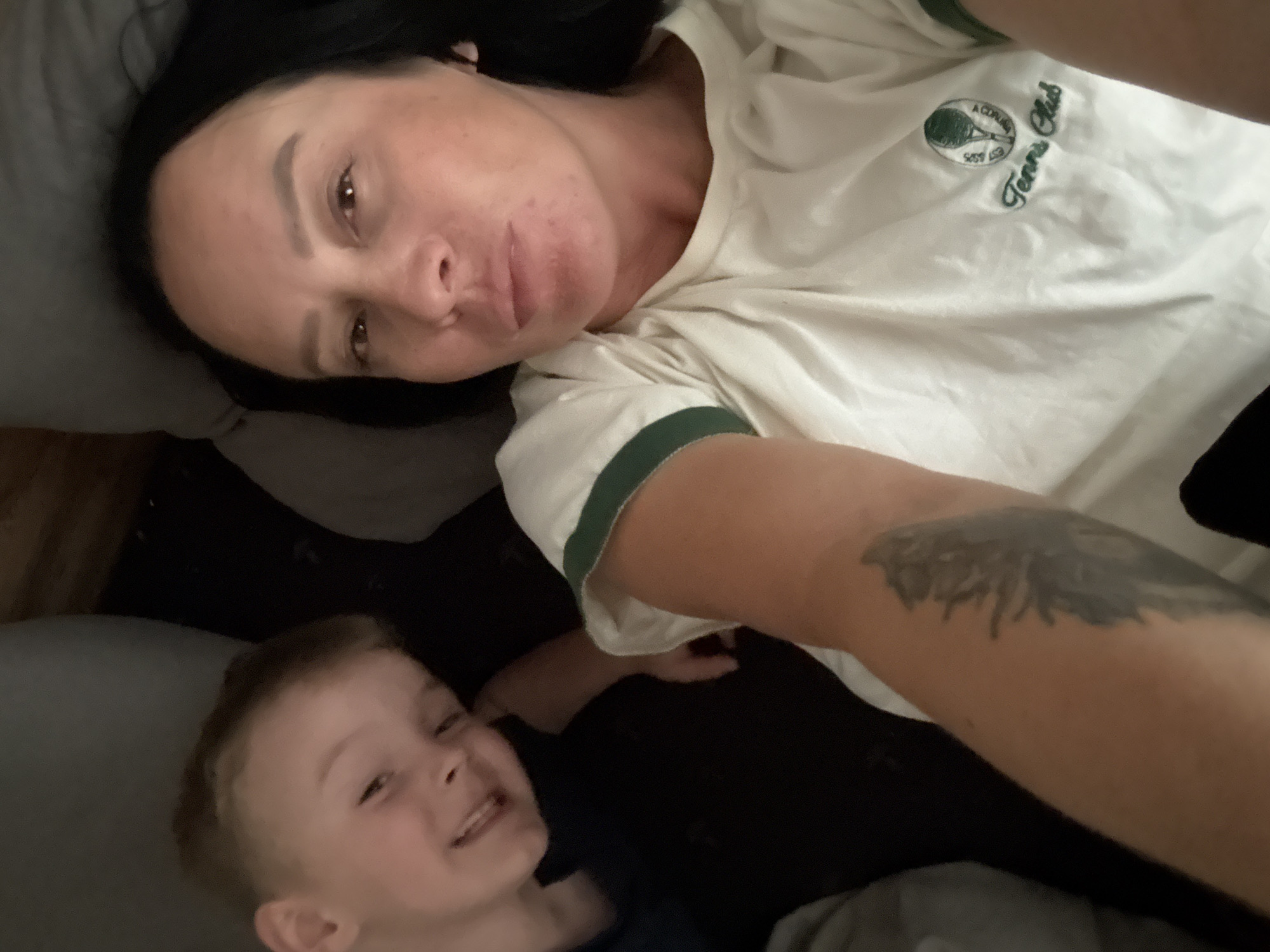 The image shows a close-up, overhead shot of two people, a woman and a young boy, lying down together. The woman is positioned on the right side of the frame, her face turned towards the camera, with her eyes open and a neutral expression. She has dark hair and appears to be wearing a light-colored t-shirt with green trim and an embroidered logo that includes a tennis racket and the text "A Coruña Tennis Club". A tattoo is visible on her forearm.
The young boy is on the left side of the frame, closer to the bottom. He is looking towards the camera with a wide smile, his eyes bright and his mouth open in laughter. He has short, light-brown hair and appears to be wearing a dark shirt.
They are lying on what appears to be a bed or couch, with pillows visible. The lighting is dim, suggesting it might be evening or a dimly lit room. The overall mood appears relaxed and intimate, with the two individuals seemingly enjoying each other's company. No specific cues about the weather or time of day are present beyond the dim lighting. The location context provided is Chaikovsky, Russia.