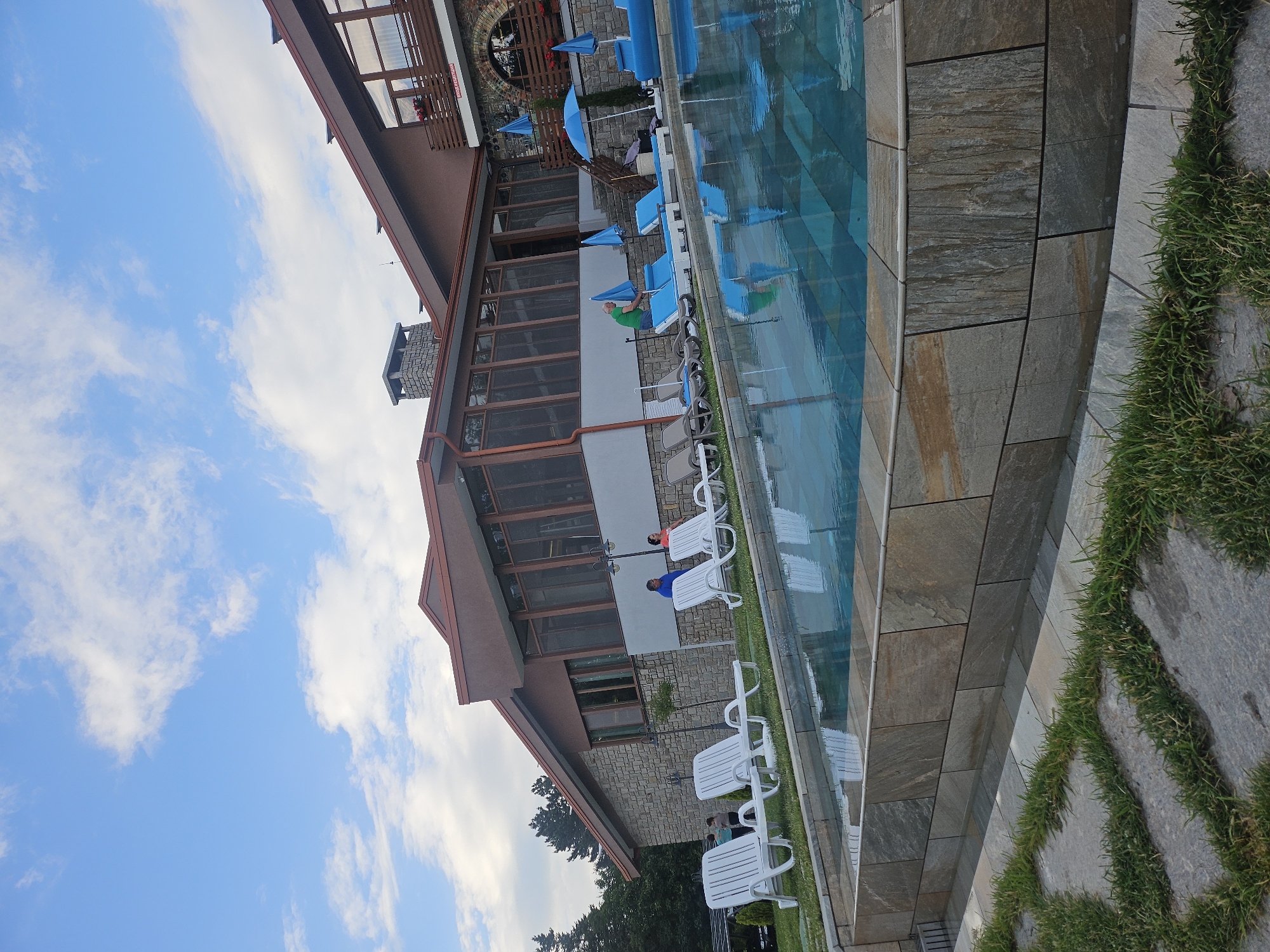 The image shows an outdoor swimming pool area adjacent to a building. The building appears to be a hotel or resort, with a rustic stone facade and a reddish-brown roof. There are several white lounge chairs arranged around the pool, with some occupied by individuals. A man in a green shirt is interacting with blue umbrellas and lounge chairs. The pool itself is filled with clear blue water. The surrounding environment includes a grassy area with stone pathways leading to the pool, and trees in the background. The sky is partly cloudy. The location is identified as Dobrostan, Bulgaria.