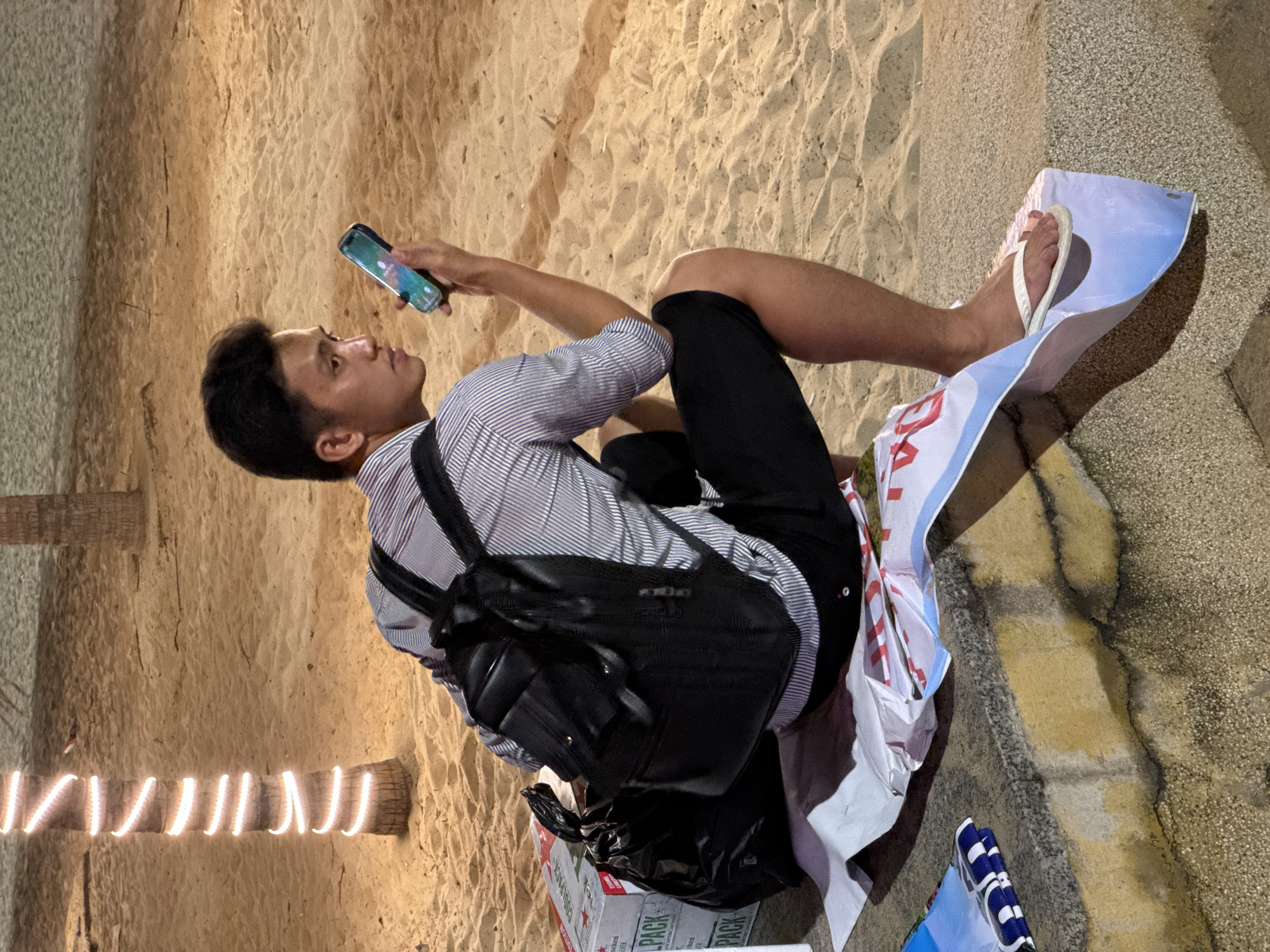 A young East Asian male wearing a striped long-sleeved shirt, black shorts, and white flip-flops sits on a concrete ledge at a beach at night. He is holding a smartphone in his right hand, looking at its screen. A black backpack is slung over his shoulders, and a black plastic bag rests on the sand next to several cardboard boxes. The sand is illuminated by what appear to be string lights wrapped around a palm tree. The background shows more sand and palm trees, suggesting a tropical location.