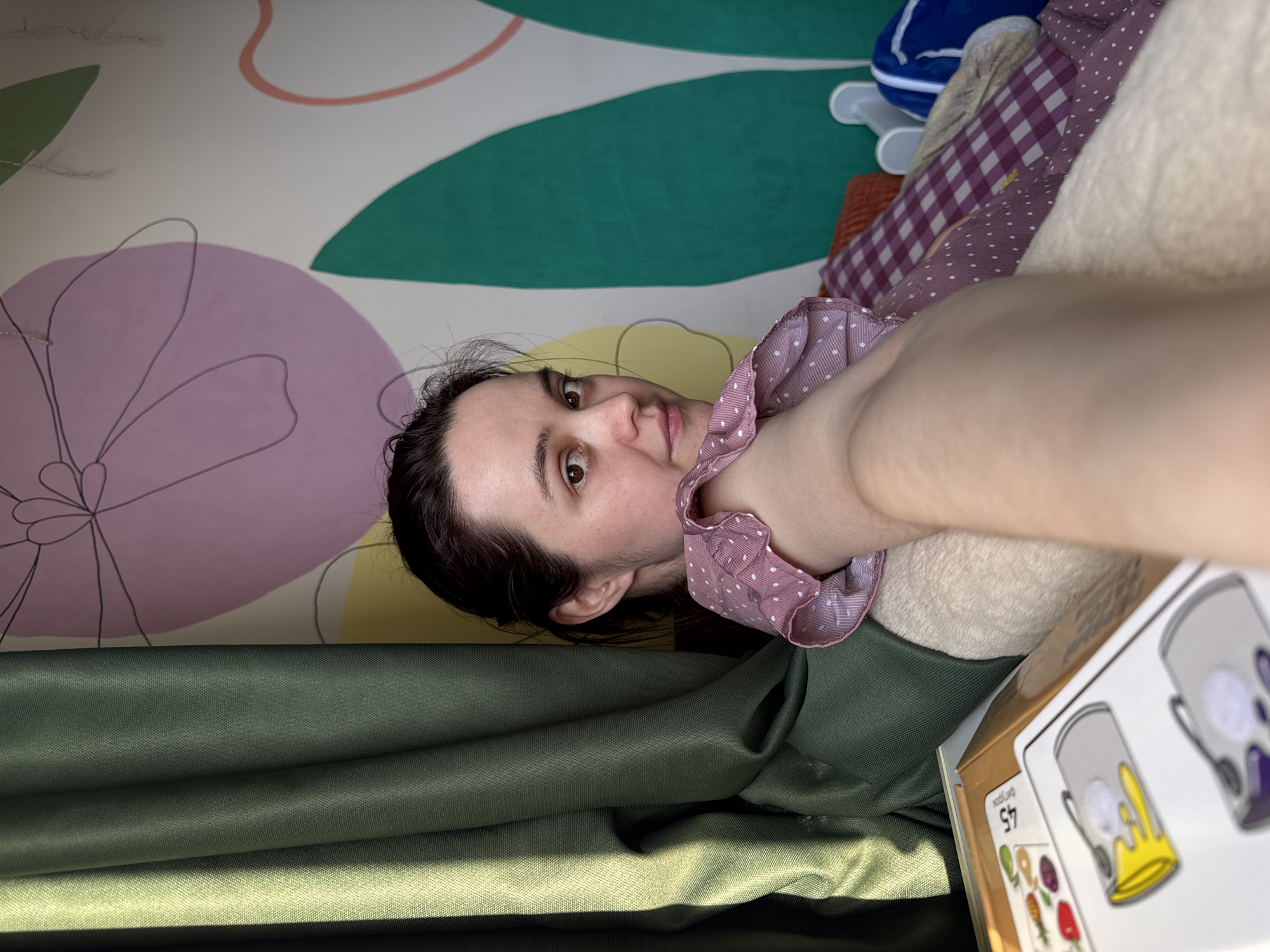 A woman with brown hair and eyes is taking a selfie. She is wearing a pink and white polka-dotted top. Her background is a wall with a colorful flower design. There is also green fabric visible in the foreground. A game box is visible near her. The location is not identifiable as being in Izhevsk, Russia from the given information.