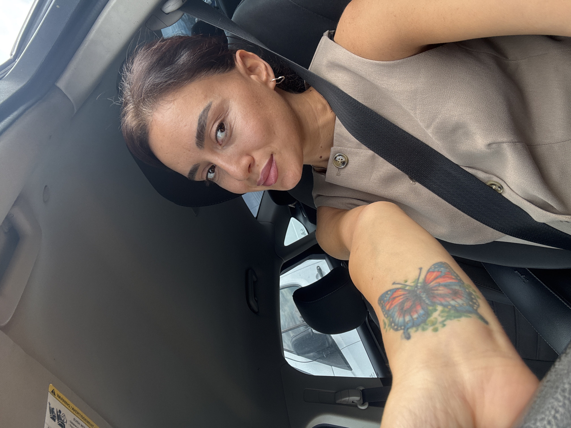 A woman is sitting in the driver's seat of a car, looking at the camera. She has her left arm extended towards the camera, displaying a butterfly tattoo. She is wearing a tan sleeveless vest with a button closure. A black seatbelt is diagonally across her chest. The interior of the car is visible, including the dashboard, sun visor, and parts of the seats. Outside the car window, another vehicle is visible. The scene appears to be a street in Tbilisi, Georgia.