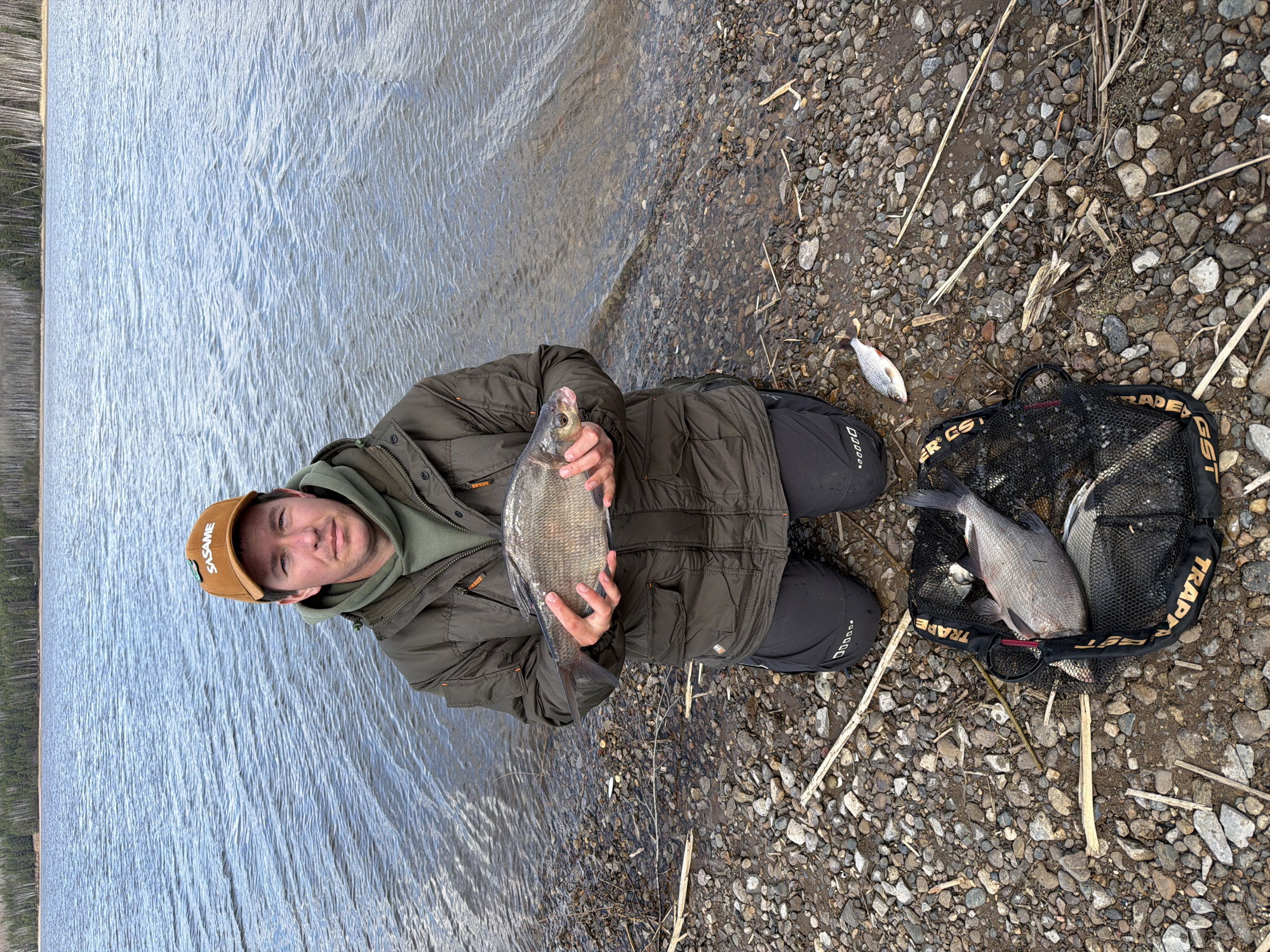 A man is captured in a portrait shot, kneeling on a pebbled shoreline beside a large body of water, likely a lake or river in Chaikovsky, Russia. He is holding a medium-sized fish, possibly a Bream, with both hands, presenting it to the camera.
The man wears a brown baseball cap with the word "SASAME" visible on the front, an olive green hooded sweatshirt, and a dark olive green quilted jacket with "URSUS" text on the shoulder. His trousers are dark, with a patterned detail (possibly "00000") on the left leg. His expression is neutral to slightly smiling, suggesting contentment with his catch.
The water surface behind him is rippled and reflects the soft, diffused light of an overcast day. In the distance, a faint treeline marks the opposite bank or horizon. The immediate foreground features a rocky and pebbly shore, interspersed with dry reeds and twigs.
To the man's right, a black mesh fishing net or keepnet is laid on the ground. Several fish, similar in size and type to the one he is holding, are visible inside the net. Another smaller, silver-colored fish lies on the pebbles outside the net. Visible text on the net includes "TRAP" and "R GS", likely part of a brand name like "TRAPER GST". The scene depicts a successful fishing outing in a natural outdoor environment.