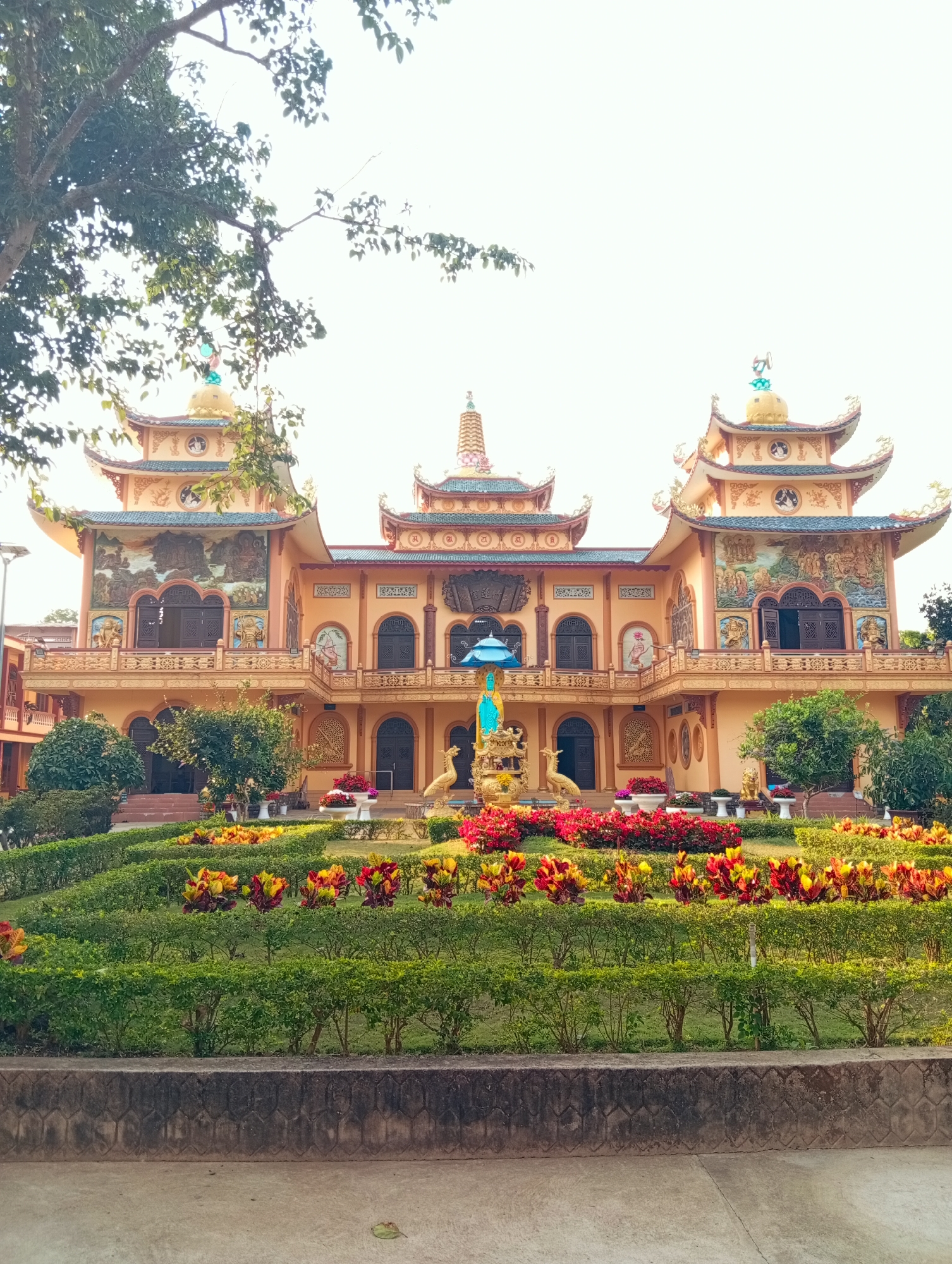 The image depicts a brightly colored Buddhist temple situated in a garden setting in Phú An, Vietnam. The temple features ornate architecture with multiple levels and intricately decorated façades. The building is predominantly painted in hues of light orange and yellow, adorned with detailed artwork and sculptures. The roofs are turquoise. Lush greenery and colorful flowerbeds surround the temple, creating a serene atmosphere. A statue of Buddha sits on a fountain. The sky appears overcast. The ground in the foreground is concrete.