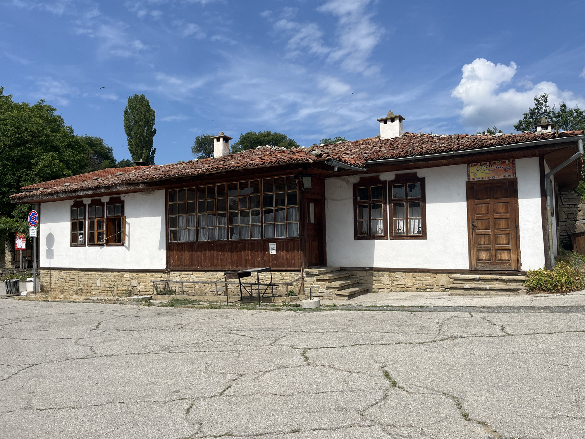A white-walled, single-story building with a red-tiled roof stands under a bright blue sky with scattered clouds. The building has several wooden-framed windows, a prominent wooden double door on the right, and a smaller entrance with a wooden door on the left. The lower portion of the building is constructed with light-colored stone blocks. In front of the building, there is a paved area with visible cracks. Two metal chairs and a small table are placed in front of the left side of the building. A sign with Cyrillic text and decorative patterns is mounted above the wooden double door. The environment appears to be a village, with trees visible in the background and a tall, slender tree to the left of the building. The building is located in Zheravna, Bulgaria.