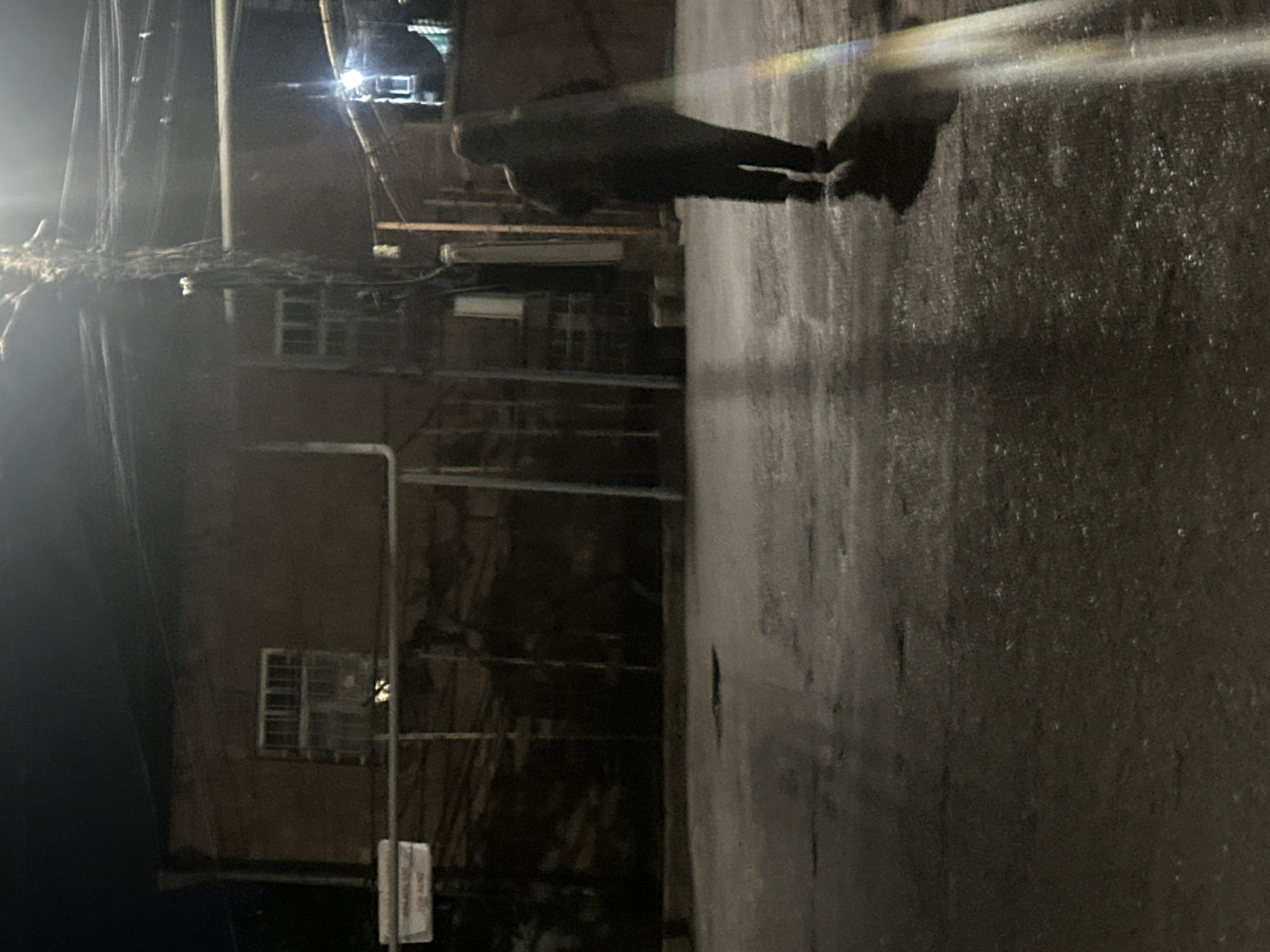 The image, captured at night, depicts a lone figure standing on a wet street or alleyway. The person, dressed in dark attire, is seen from behind and slightly to the side, appearing as a silhouette against the reflective ground. They are looking towards the left side of the frame, where dark, unassuming buildings with multiple windows line the path. Overhead and along the building facades, a dense network of utility wires, pipes, and cables is visible.
The ground surface, glistening with moisture likely from recent rain, reflects the artificial light sources present. Several bright lights, possibly streetlights, are visible in the upper left, causing prominent lens flares and a distinct, rainbow-hued streak across the wet pavement. Another light source is dimly seen in the upper background. The overall scene is characterized by deep shadows and muted tones of gray, black, and brown, with bright white and colored light accents. A small, rectangular white sign is partially visible on the right side of the image, near the bottom, but any text on it is unreadable due to blurriness. The scene conveys a quiet, possibly melancholic, urban night setting.
