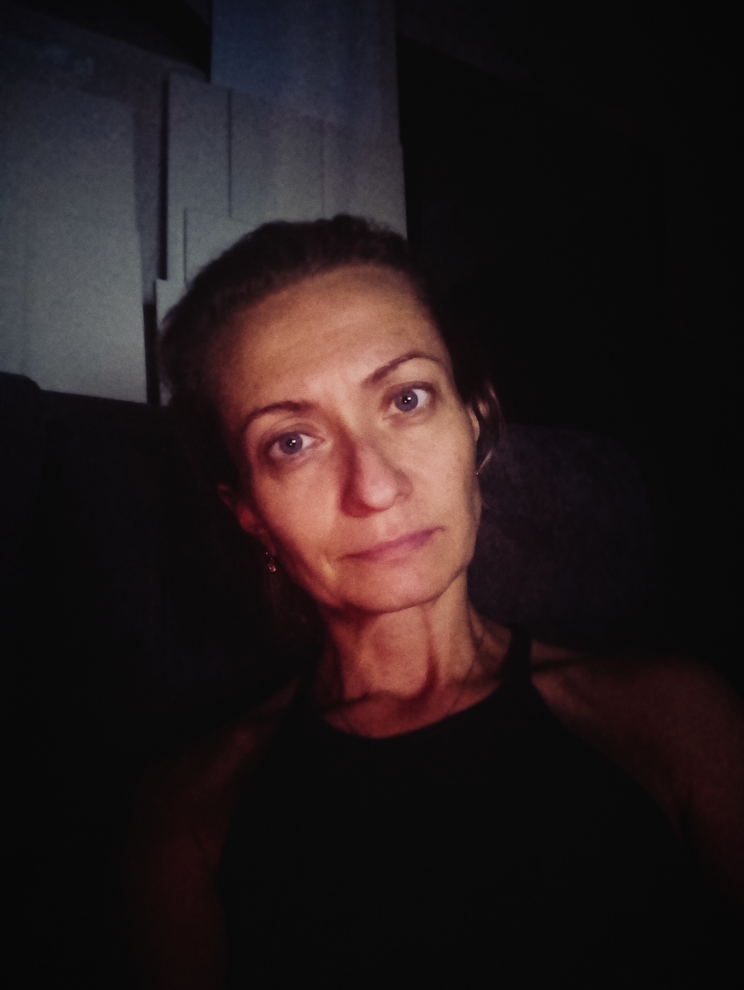 The image is a low-light, close-up selfie of a woman with fair skin, light brown hair pulled back, and blue eyes. She is wearing a dark, sleeveless top. Her face is illuminated from the front, creating shadows on the right side of her face and neck. In the background, to her left, is a dark, textured wall or partition. The overall impression is one of intimacy and perhaps a dimly lit interior space. There is no discernible indication of the specific location in Moscow, Russia, or any other urban detail.