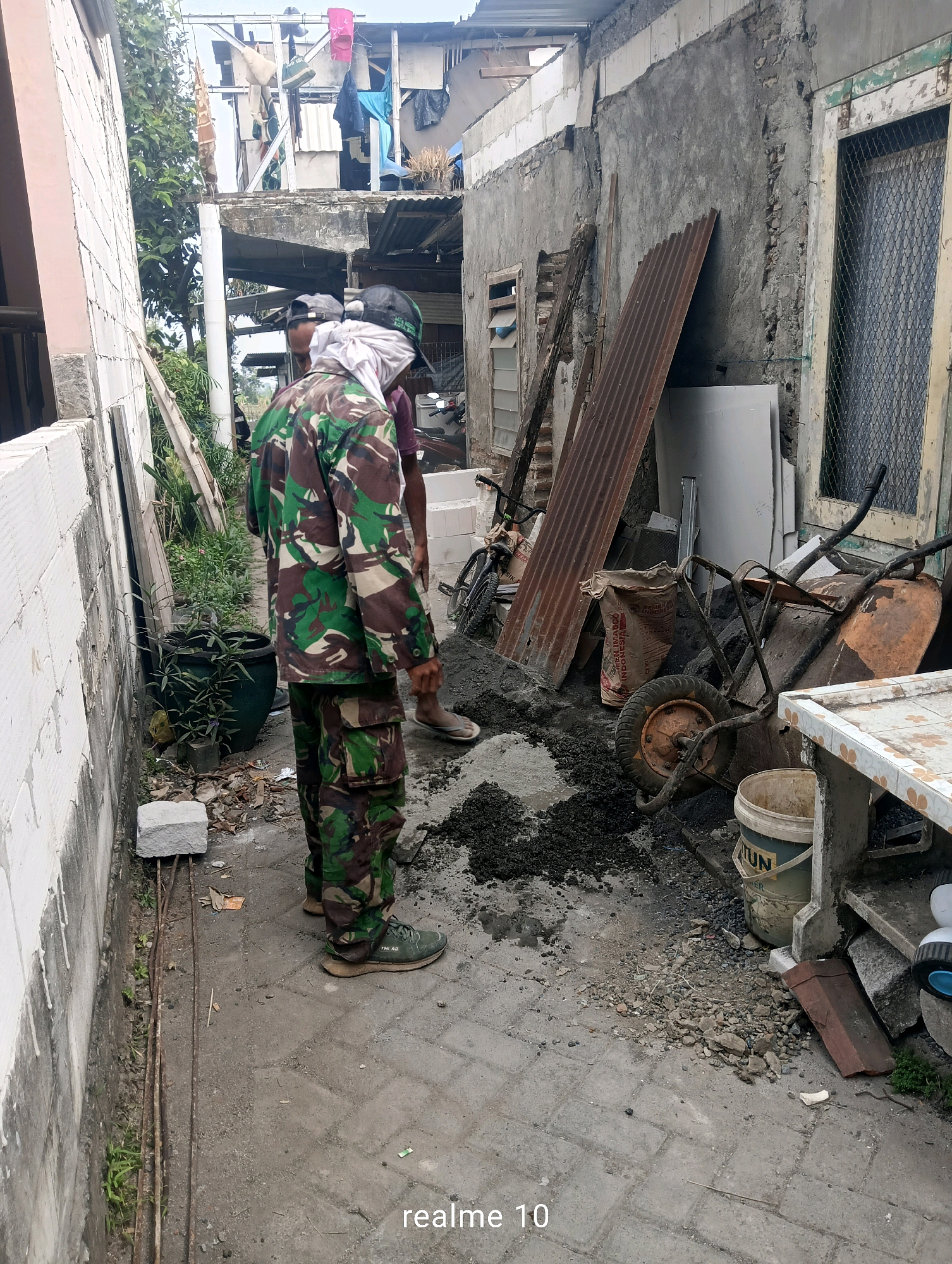 In Batu, Indonesia, two individuals in camouflage clothing are present in a narrow alleyway. One individual, on the left, is partially obscured, wearing a camouflage jacket and pants, and appears to be holding a white cloth around their head. The other individual, on the right, is also wearing camouflage attire. They are standing near a pile of dark material, likely cement or wet soil, and a wheelbarrow. The environment is characterized by a mix of old and new construction, with concrete walls, a corrugated metal sheet leaning against a wall, and various building materials scattered around. Laundry is visible hanging from clotheslines above, suggesting a residential area. The ground is paved with interlocking concrete blocks. The overall scene depicts a moment of construction or maintenance activity in a somewhat dilapidated urban setting.