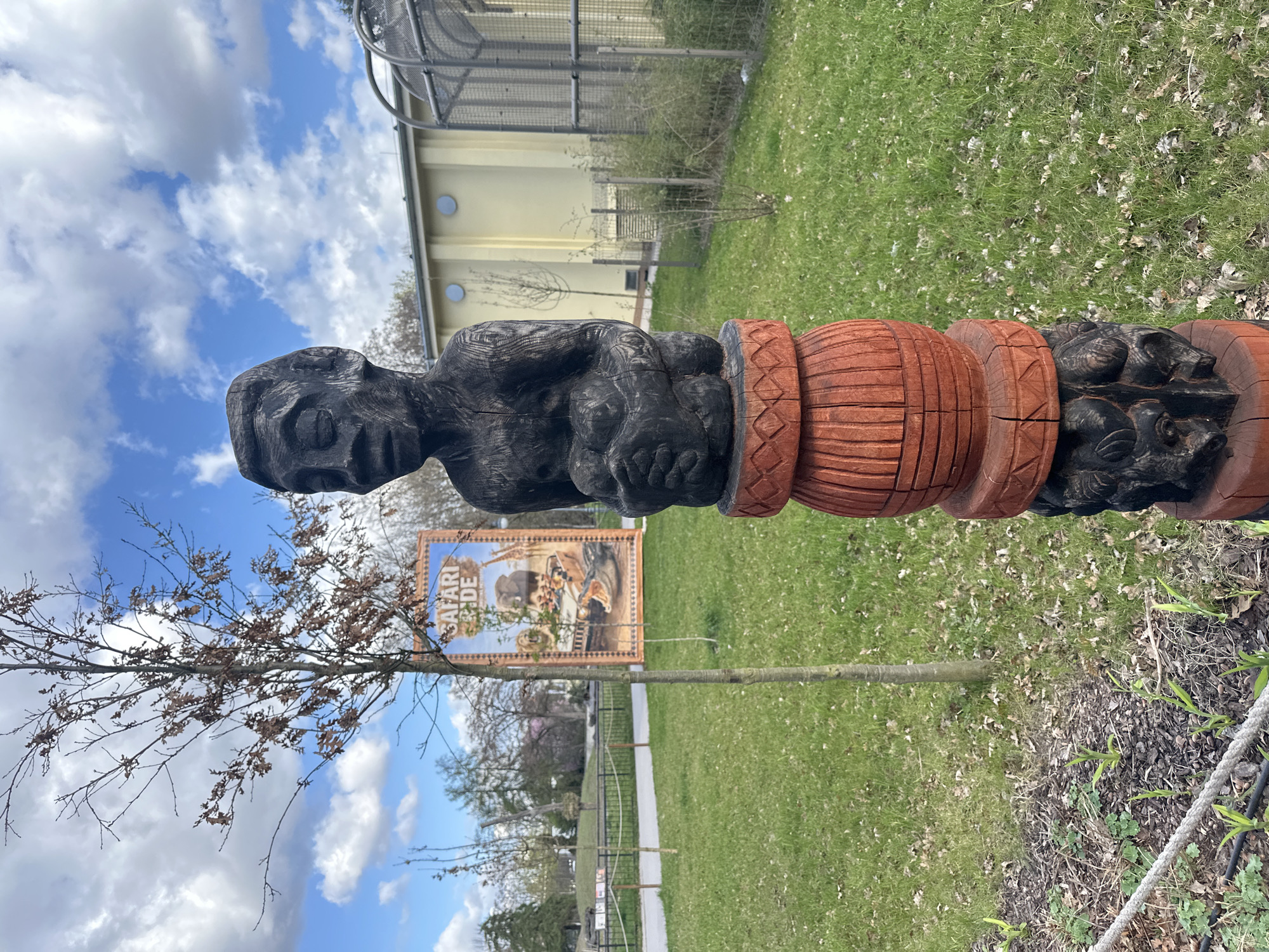 A carved wooden totem pole depicting a figure is located in a green area. The statue has a brown base, a black torso, and a black head. Behind the totem pole is a sign with the words "SAFARI" and an image of various animals. The scene is situated in Wrocław, Poland. A building and a partially cloudy sky are visible in the background. There is a young tree planted near the totem pole. A rope fence is visible in the lower right corner of the image.