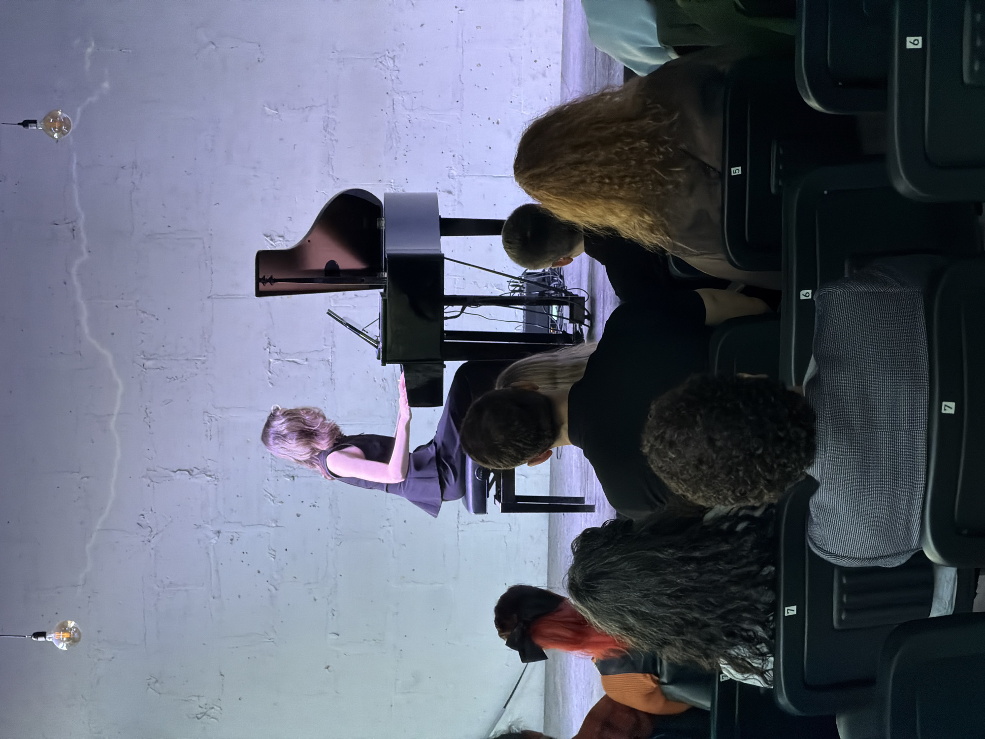 The scene depicts a woman playing a piano on a stage in front of an audience. The audience members are seated in rows of chairs numbered 5, 6, and 7. The room appears to be indoors with white brick walls and exposed light bulbs hanging from the ceiling. The location is in the city of Unknown, Unknown.