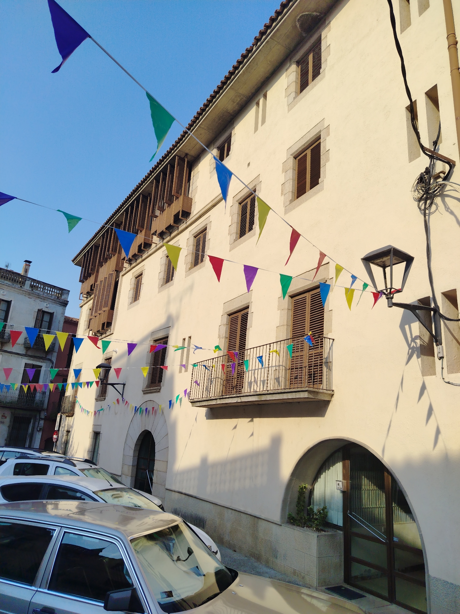 The image captures a street scene in the city of Hostalric, Spain, likely during a festive occasion. The focus is on a pale yellow building adorned with strings of colorful triangular flags, which stretch across the street and are draped along the facade. The building features several windows with wooden shutters, and a balcony with a decorative railing is visible on the upper floor. Below the balcony, a large arched doorway leads into the building.
In the foreground, several parked cars are visible, including a light-colored sedan and a white SUV. The street appears to be paved, with a slight incline. The sky above is a clear blue, and the sunlight casts shadows of the flags and the building onto the wall, suggesting it is daytime. A street lamp is mounted on the building's facade, near the balcony. The architecture suggests a historic European setting. No people are visible in the frame.