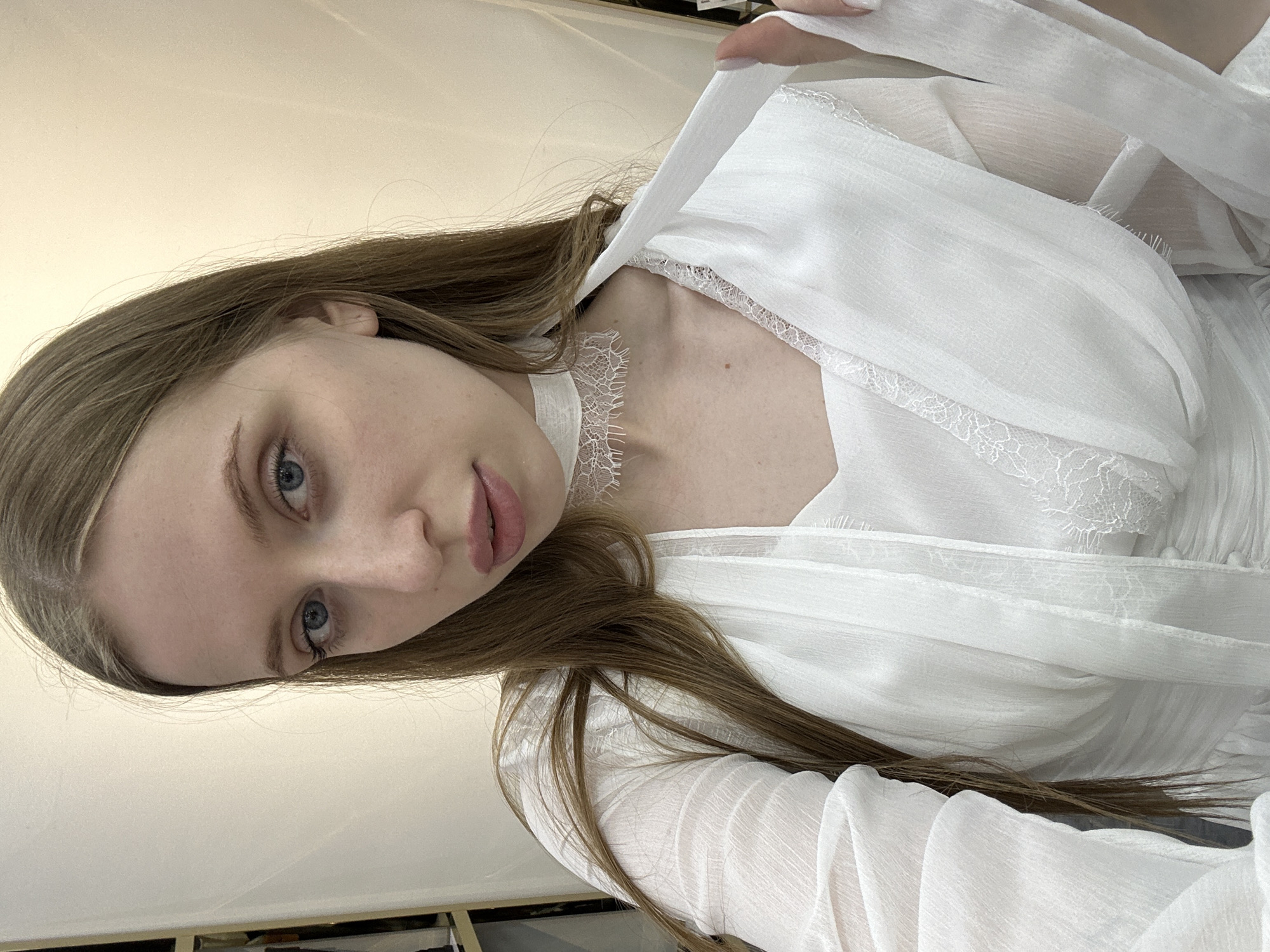 A fair-skinned, light-haired woman is wearing a white, lacy garment. The image appears to be taken from a high angle, capturing her face and upper torso. The background is out of focus, but suggests an indoor location, possibly in Chaikovsky, Russia.