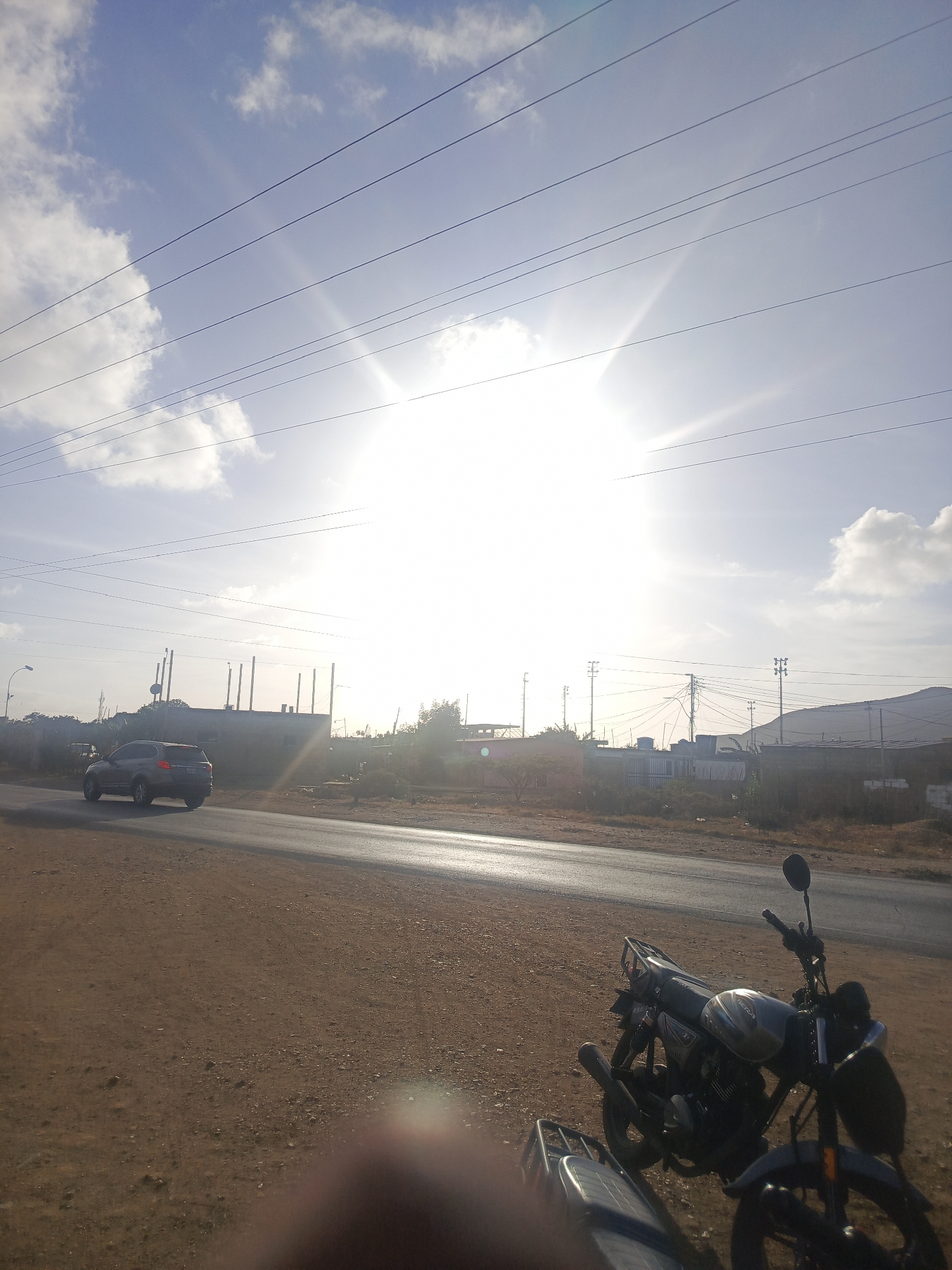 The image shows an outdoor scene in Juan Griego, Venezuela. A paved road runs horizontally across the middle of the frame. A gray SUV is driving on the road to the left. Buildings and structures are visible in the background, including utility poles. Overhead power lines run across the sky. Two motorcycles are parked on the dirt shoulder of the road in the foreground on the right. A blurry dark object is in the bottom center, obstructing the full view. The sky is light blue with scattered clouds, and the sun is brightly shining.
