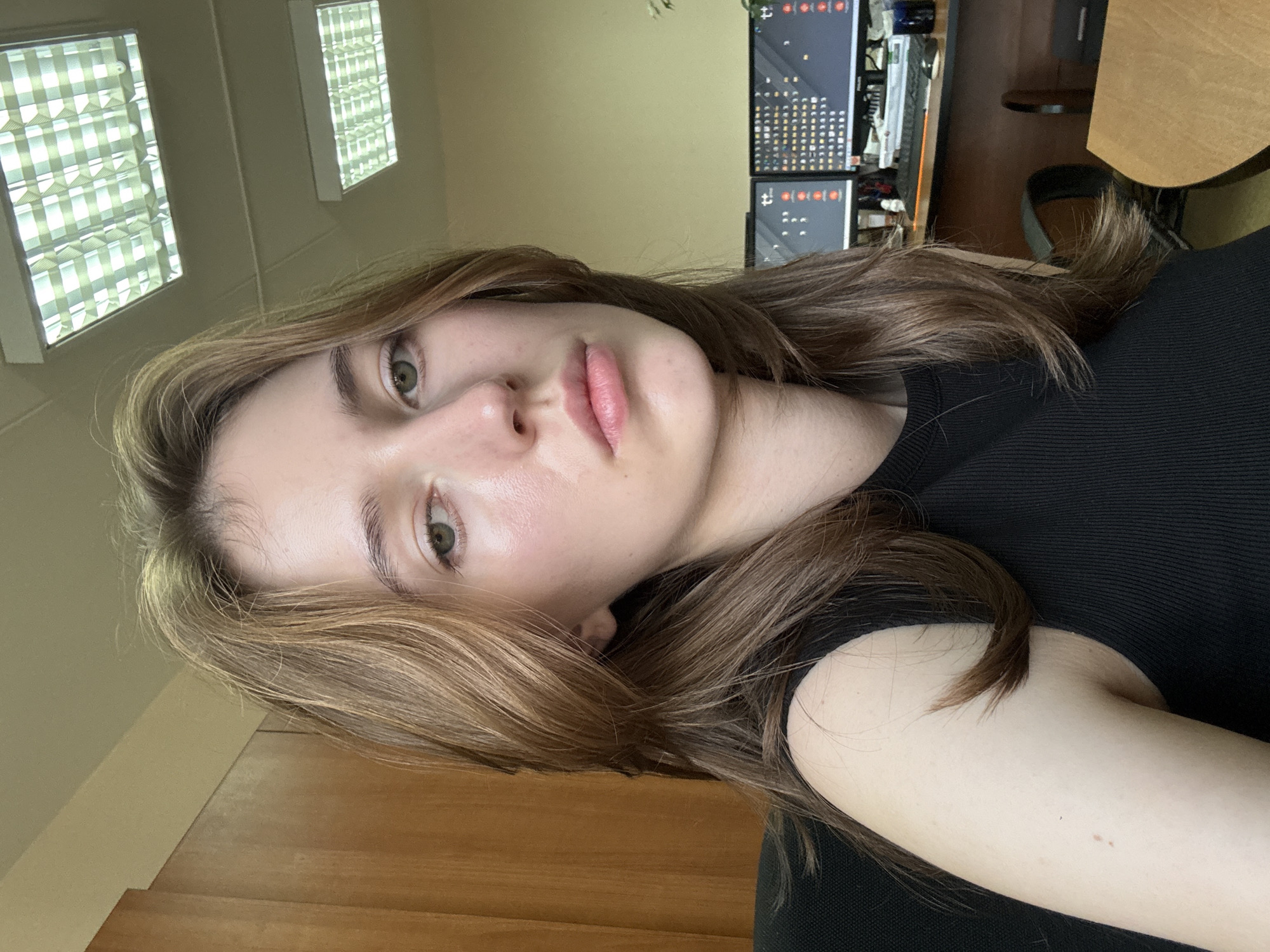 A young Caucasian woman with shoulder-length brown hair is looking towards the camera. She is wearing a black sleeveless top. Her skin appears pale and somewhat flushed on her cheeks. Her lips are slightly parted. She is seated at a desk in what appears to be an office environment. Behind her, two computer monitors are visible, displaying what looks like code or data. The room is lit by fluorescent ceiling lights. The overall ambiance suggests an indoor setting, likely a workspace, with a desk and office furniture. The specific location of Perm, Russia, cannot be determined from the visual information provided in the image.