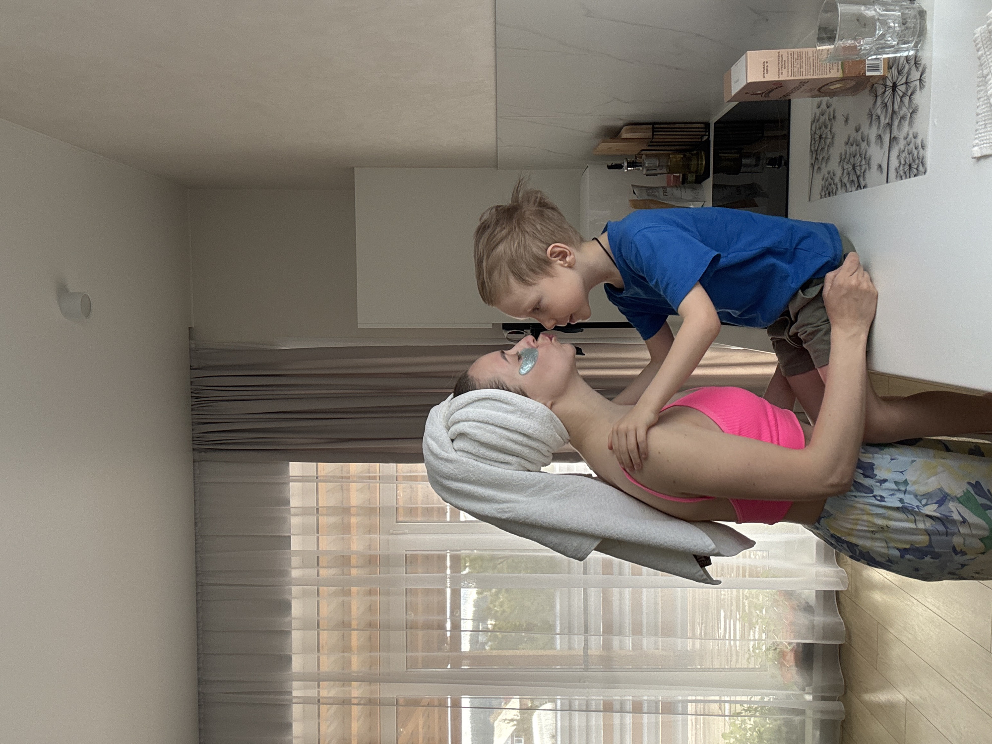 The image depicts a domestic scene inside a house in Chaikovsky, Russia. A woman wearing eye patches, a pink top, and floral-patterned shorts lies on a white counter. A light gray towel is wrapped around her head. A young boy wearing a blue t-shirt and khaki shorts leans over her, looking at her face and touching her neck area with his hands. A window with closed blinds is visible in the background. A cabinet is on the right side of the image, holding various items including food boxes and glassware.