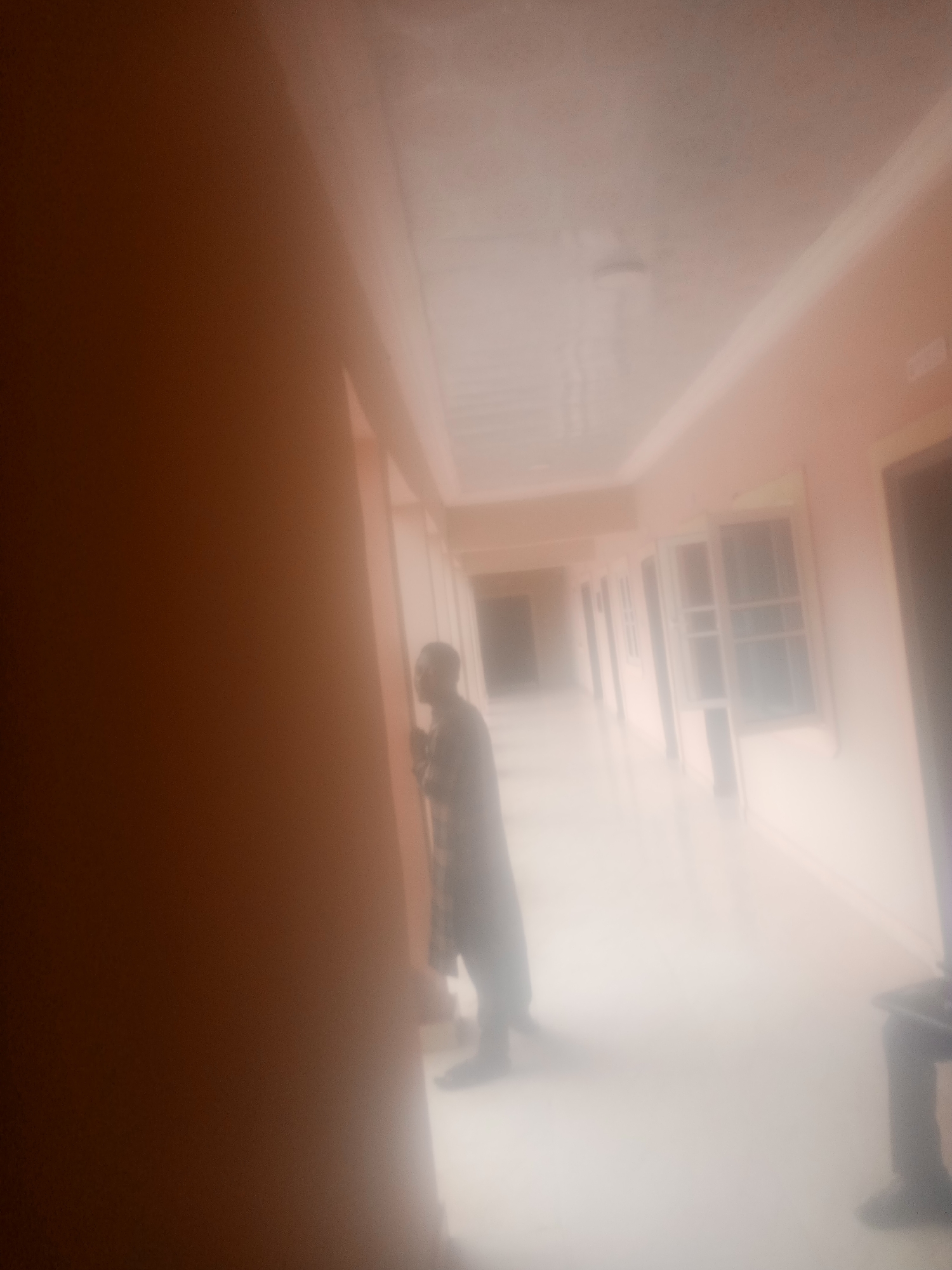 The image depicts a well-lit, indoor hallway with a person standing on the left side, facing away from the camera and looking into the hallway. The hallway appears to be a residential or institutional building, possibly a dormitory or hotel, with multiple doors or windows visible along the right wall. The walls are painted a light peach color, and the ceiling is white with fluorescent lighting. The floor is a light color, reflecting the light from above. The subject is wearing a patterned robe and appears to be a man. The hallway extends into the distance, creating a sense of depth and perspective. The lighting is bright, suggesting it could be daytime or that the interior lighting is very effective. There is no visible text in the image. The overall atmosphere is neutral and ordinary, with no strong indications of a specific event or emotion.