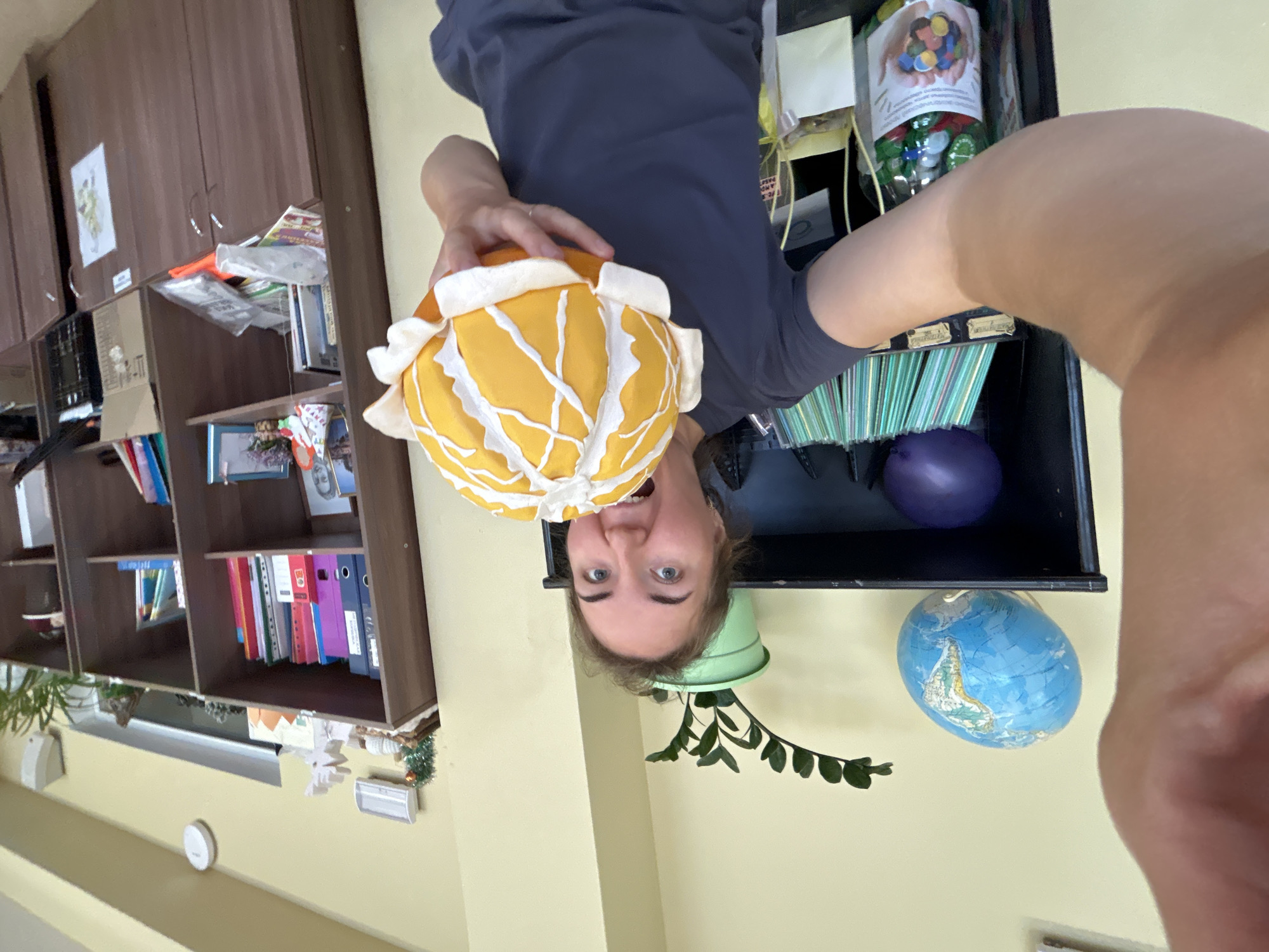 An indoor, eye-level shot captures a young adult, likely female, with an expressive face, holding a spherical yellow and white object. Her head is tilted slightly to the side, eyes wide, and mouth open in a joyful or surprised expression, indicating playfulness or excitement. She wears a dark grey or navy t-shirt. The yellow and white object she holds resembles a stylized, segmented fruit like a peeled orange or a flower.
The setting appears to be a room, possibly an office, classroom, or study, with light yellow/cream walls. In the background, on the left, is a brown wooden bookcase filled with various items, including books, colorful binders (red, blue, purple, green), papers, and a cardboard box. A small potted plant with green leaves is also visible on a lower shelf of this unit.
On the right side of the frame, another shelving unit or cubbyhole is visible. The top shelf holds a clear container labeled "СУВЕНИРЫ" (Souvenirs) with an image of hands cupping colorful objects and text below that reads "С ДУШОЙ И ЛЮБОВЬЮ" (with soul and love) or similar. Below this, there's a collection of green and white straws, a small purple spherical object, a green potted plant with leaves trailing downwards, and a small blue globe resting on a shelf. The overall impression is one of a vibrant, organized, yet personal space. The photo itself is oriented sideways, giving the impression of the subject being upside down, which enhances the playful mood.