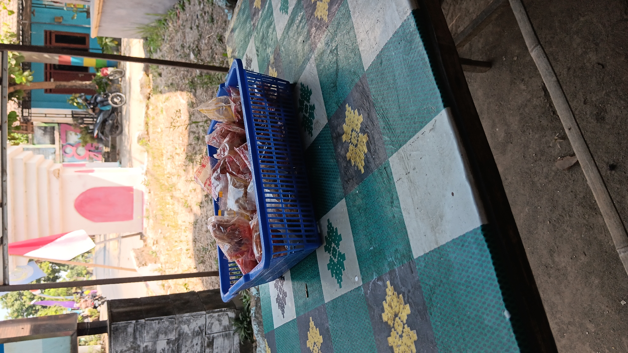 A blue plastic basket filled with bagged snacks is placed on a table covered with a checkered tablecloth. The tablecloth has a pattern of green, white, and grey squares with yellow floral designs. In the background, a red and white flag is visible, along with a white archway structure. There are also motorcycles and buildings in the distance, suggesting an outdoor location in a residential or commercial area, likely Kelurahan, Indonesia.