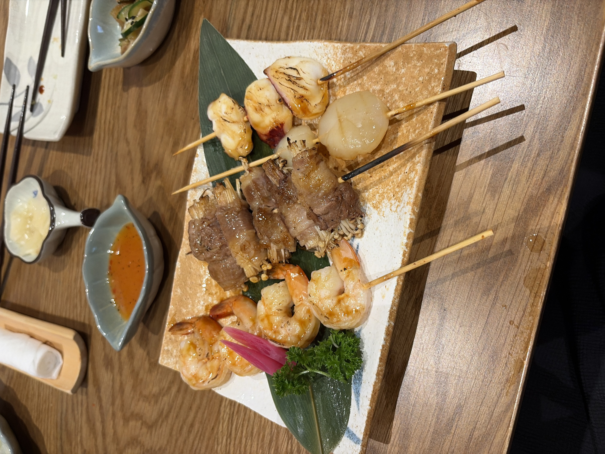 This image captures a close-up, elevated perspective of a meal served on a polished wooden table in Thủ Đức, Vietnam. The central focus is a rectangular, textured ceramic plate displaying a variety of grilled skewers. On the left side of the plate, five cooked, peeled shrimp with tails intact exhibit a golden-brown char; one shrimp is secured on a wooden skewer. Accompanying the shrimp are a small garnish of curly green parsley and two vibrant magenta-colored pickled ginger slices. Adjacent to these, four skewers of meat-wrapped enoki mushrooms are arranged, displaying a glossy, browned surface and some charring. To the right, four skewers hold grilled scallops, characterized by their firm, white flesh and partially seared, caramelized exteriors.
Surrounding the main plate, several smaller ceramic dishes are visible. In the upper left, a small light blue-green bowl contains a creamy yellow substance. To its right, a leaf-shaped light blue-green bowl holds an orange-red dipping sauce. Further to the right, a small white bowl contains a clear liquid with dark specks. In the upper right corner, another light blue-green bowl contains sliced cucumbers garnished with sesame seeds.
A light wooden tray holding a rolled white cloth is positioned in the upper center. A white rectangular plate with grey leaf patterns, bearing residual food marks and with dark chopsticks resting on it, is partially visible in the upper right quadrant. The wooden skewers from the main plate extend onto the table, casting subtle shadows. The table surface itself is a medium-brown wood with visible grain and a smooth finish, showing a few small liquid droplets below the main serving plate. No individuals are visible in the scene. The lighting suggests an indoor setting, likely a restaurant.