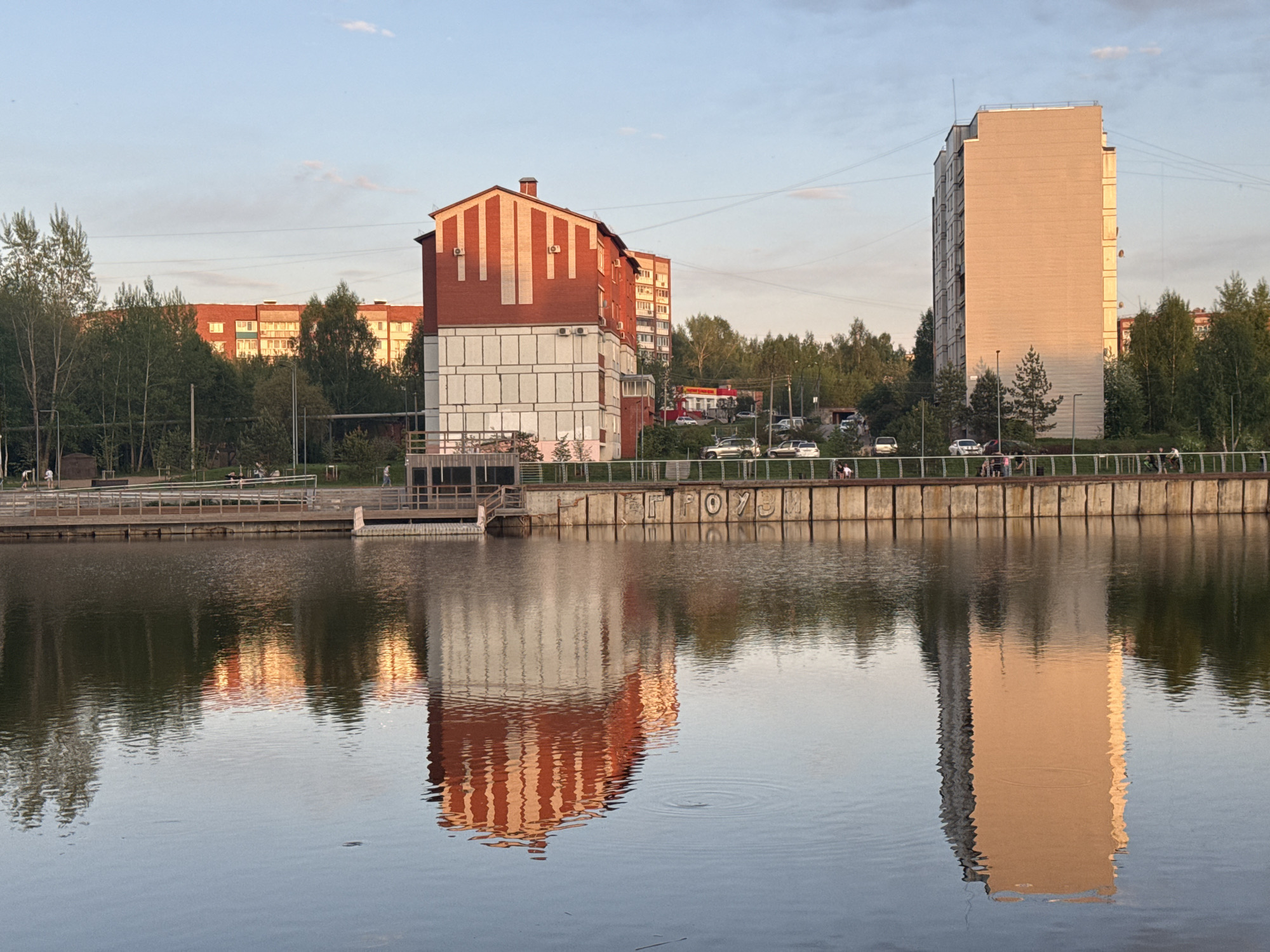 The image captures a view of Votkinsk, Russia, showcasing a calm body of water reflecting buildings and the surrounding environment. A red and white building stands to the left, mirrored in the water. To the right, a taller, gray building also has its reflection visible in the water. The backdrop consists of trees and other buildings, indicating an urban area. A concrete embankment with Cyrillic letters (ГРОУЗИМ) runs along the water's edge in the center of the image, and a few people are seen walking on the embankment. The sky appears partially cloudy, suggesting either early morning or late afternoon lighting conditions.