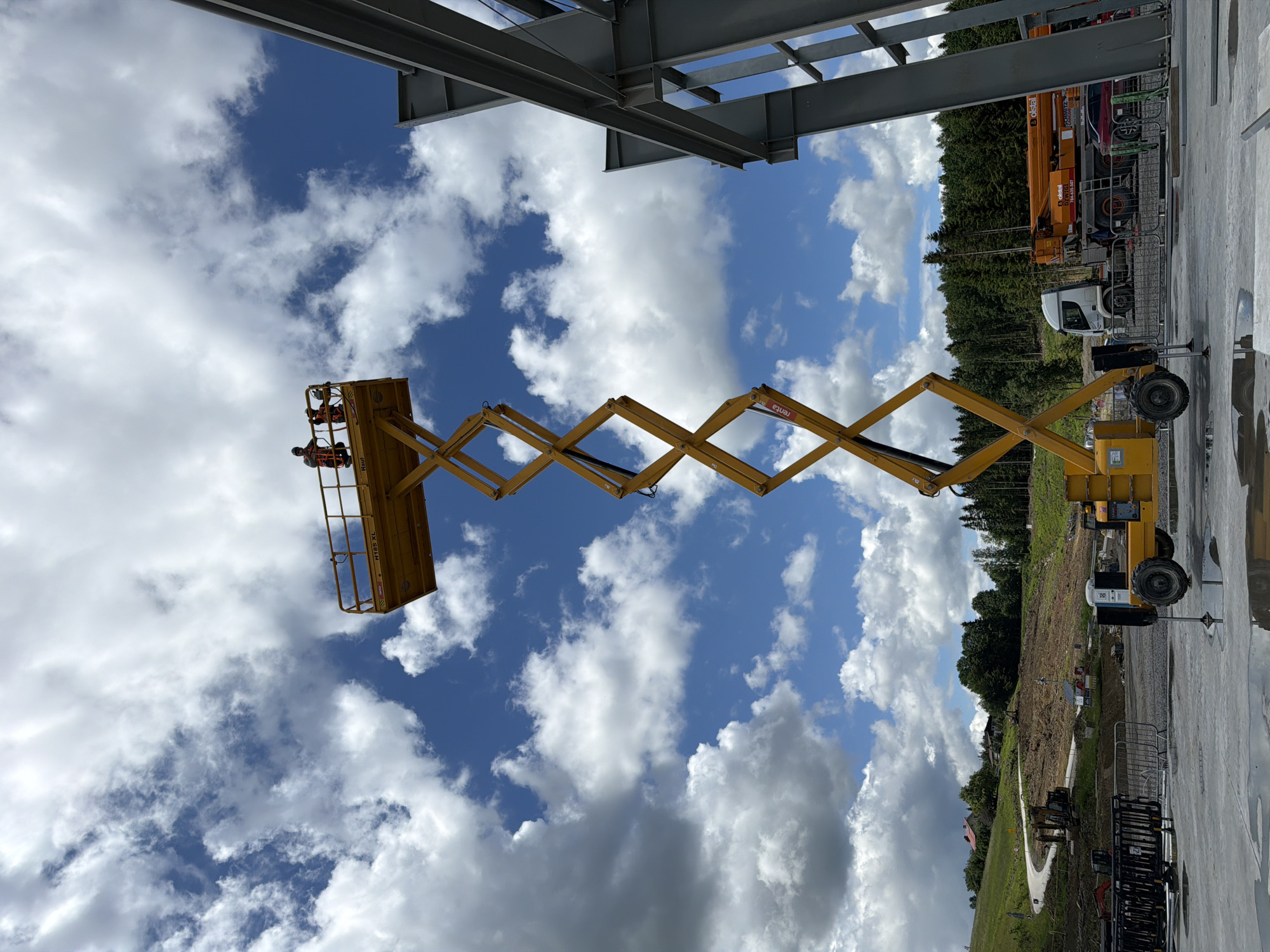 The image depicts a construction site under a cloudy sky. A tall, yellow scissor lift dominates the center of the frame, its platform elevated high in the air. Two construction workers, wearing high-visibility vests, are standing on the platform of the scissor lift. One worker is operating the machinery, while the other appears to be observing or assisting. To the right of the scissor lift, a partially constructed steel building frame is visible, suggesting ongoing construction work. In the background, a wooded hillside with a winding road and scattered houses can be seen, indicative of a rural or semi-rural setting outside of a densely populated urban area. A truck with an extended boom is parked behind a barrier, and other construction equipment, including excavators and barriers, are present on the ground. The presence of construction equipment, workers, and unfinished structures clearly indicates an active construction site.