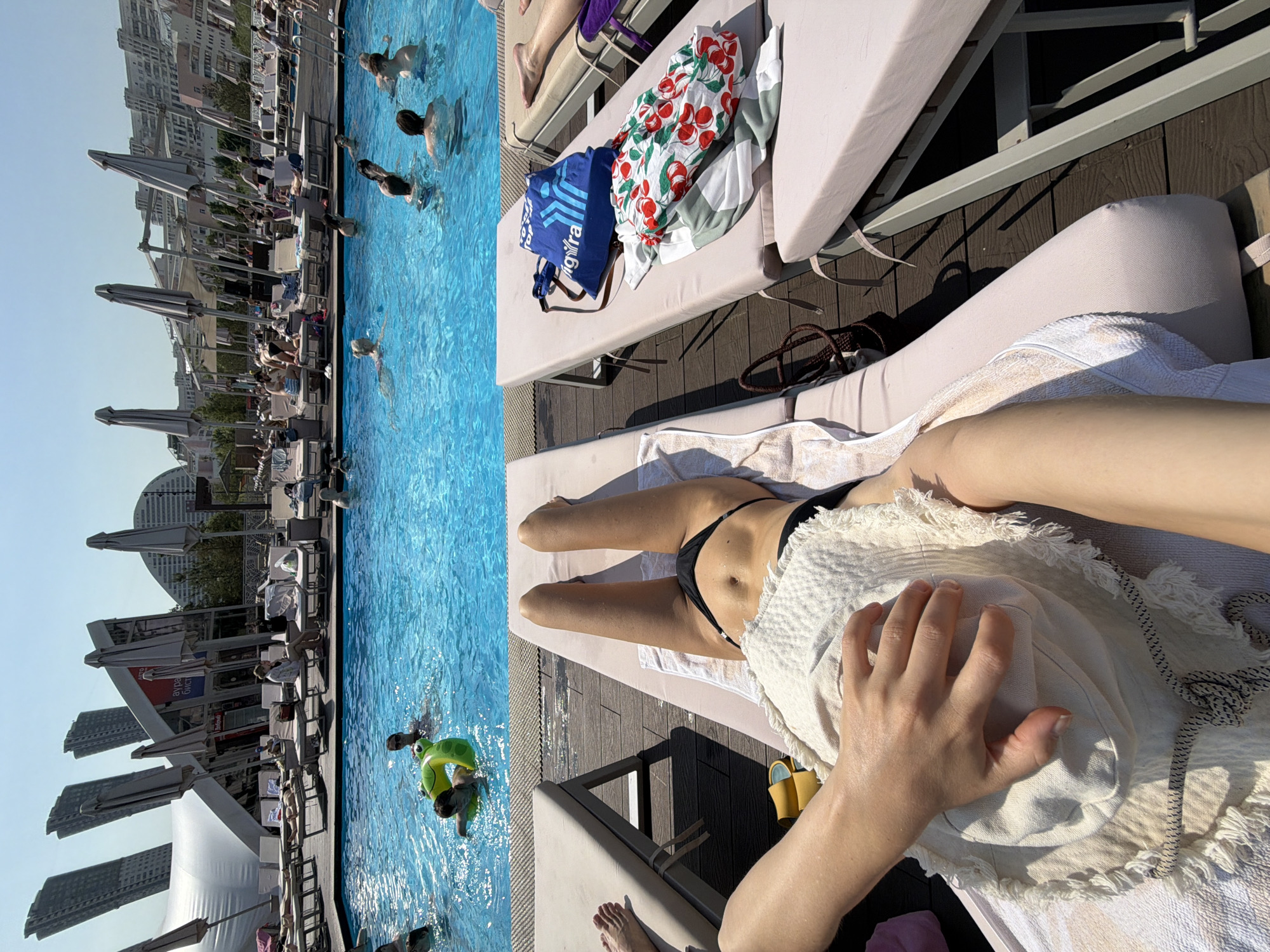The image shows a sun-drenched outdoor swimming pool area, likely a rooftop or a resort, with people swimming and lounging. In the foreground, a person is lying on a chaise lounge, holding a straw hat. Adjacent to them is another chaise lounge with beach items. In the background, there's a large blue swimming pool filled with people, some of whom are swimming or playing with a green inflatable toy. The area is lined with more chaise lounges and umbrellas, and beyond that, a cityscape featuring modern, distinctive skyscrapers and buildings is visible. The architecture and the presence of such a facility suggest a location within Moscow, Russia, possibly a well-known entertainment or recreational complex.