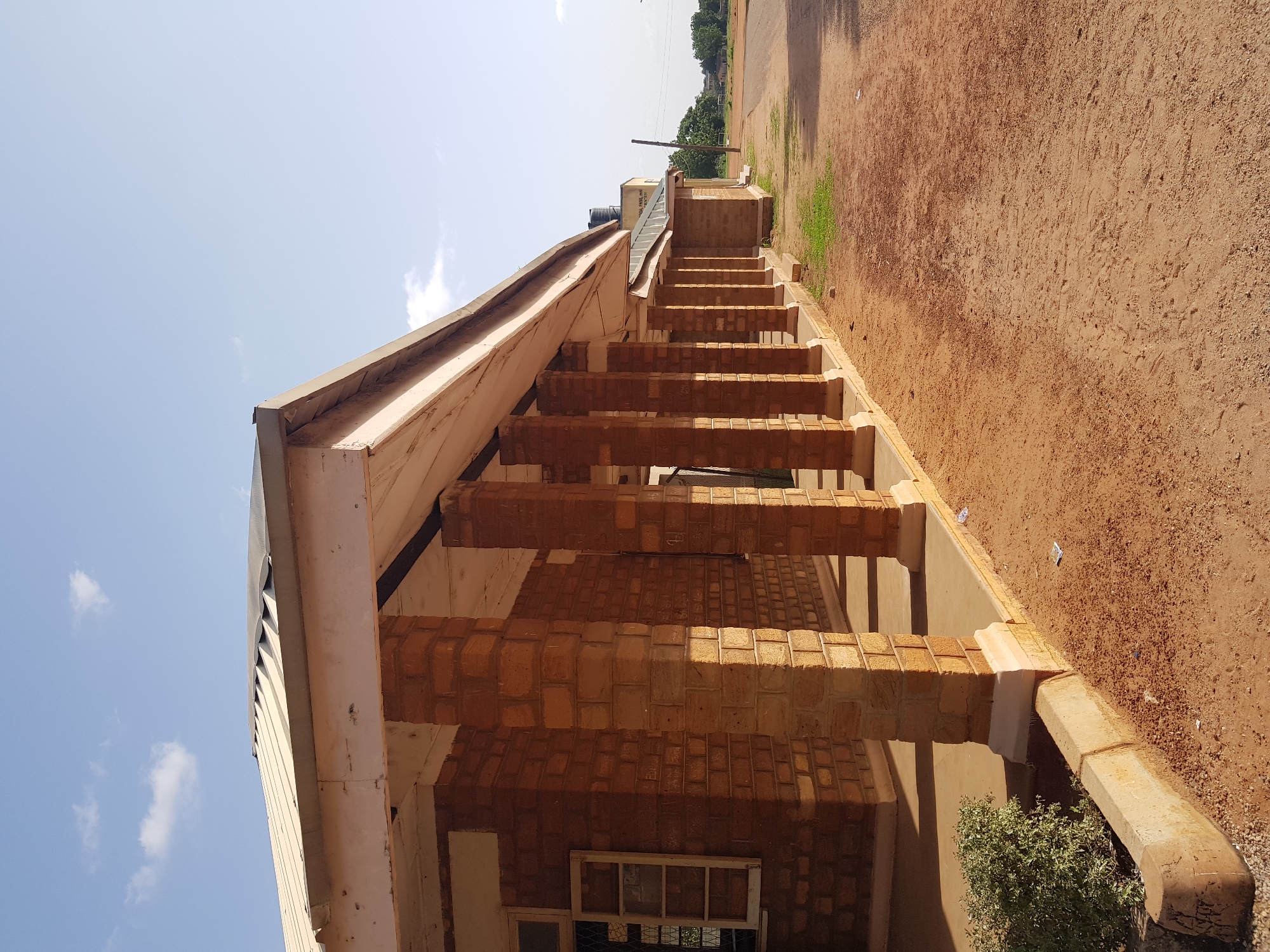 The image captures an outdoor scene in Jimeta, Nigeria, on a sunny day with a clear blue sky dotted with a few white clouds. The perspective is from a low angle, looking up at a building under construction or recently built, with a prominent staircase leading up to an entrance. The building appears to be made of light-colored bricks, with concrete elements for the stairs and supporting pillars. A large water tank is visible on the roof in the background, suggesting residential or institutional use.
The ground surrounding the building is covered in reddish-brown soil, common in many parts of Nigeria. Some sparse green vegetation grows near the base of the building. The overall impression is one of a warm, dry climate. There is no visible activity or people in the frame, giving the scene a sense of stillness. The time of day appears to be late morning or early afternoon, judging by the angle of the sunlight casting shadows.
A partially visible sign on the building in the background contains some text, but it is too blurry to read clearly. The architectural style suggests a functional building, possibly a school, clinic, or community center. The sunlight and clear sky indicate fair weather.