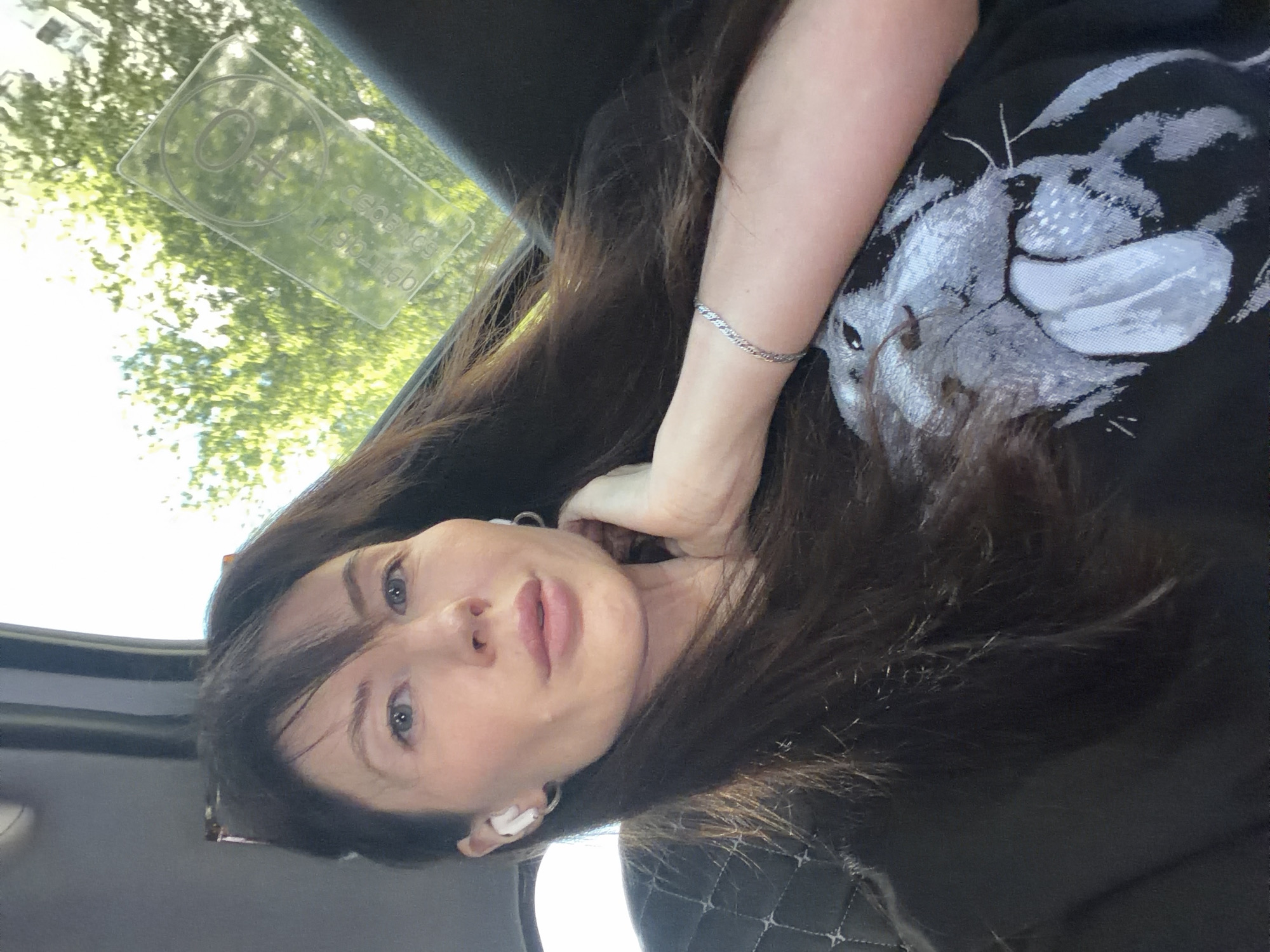 A selfie-style photograph depicts a woman inside a vehicle. She has long dark hair and is wearing a black t-shirt with a grey animal print. She is also wearing a silver bracelet on her left wrist. An airpod is visible in her left ear. The car window shows trees and a reflective sticker is adhered to the window. The seat appears to be quilted leather. Based on the language on the sticker in the window, the location is likely Perm, Russia.