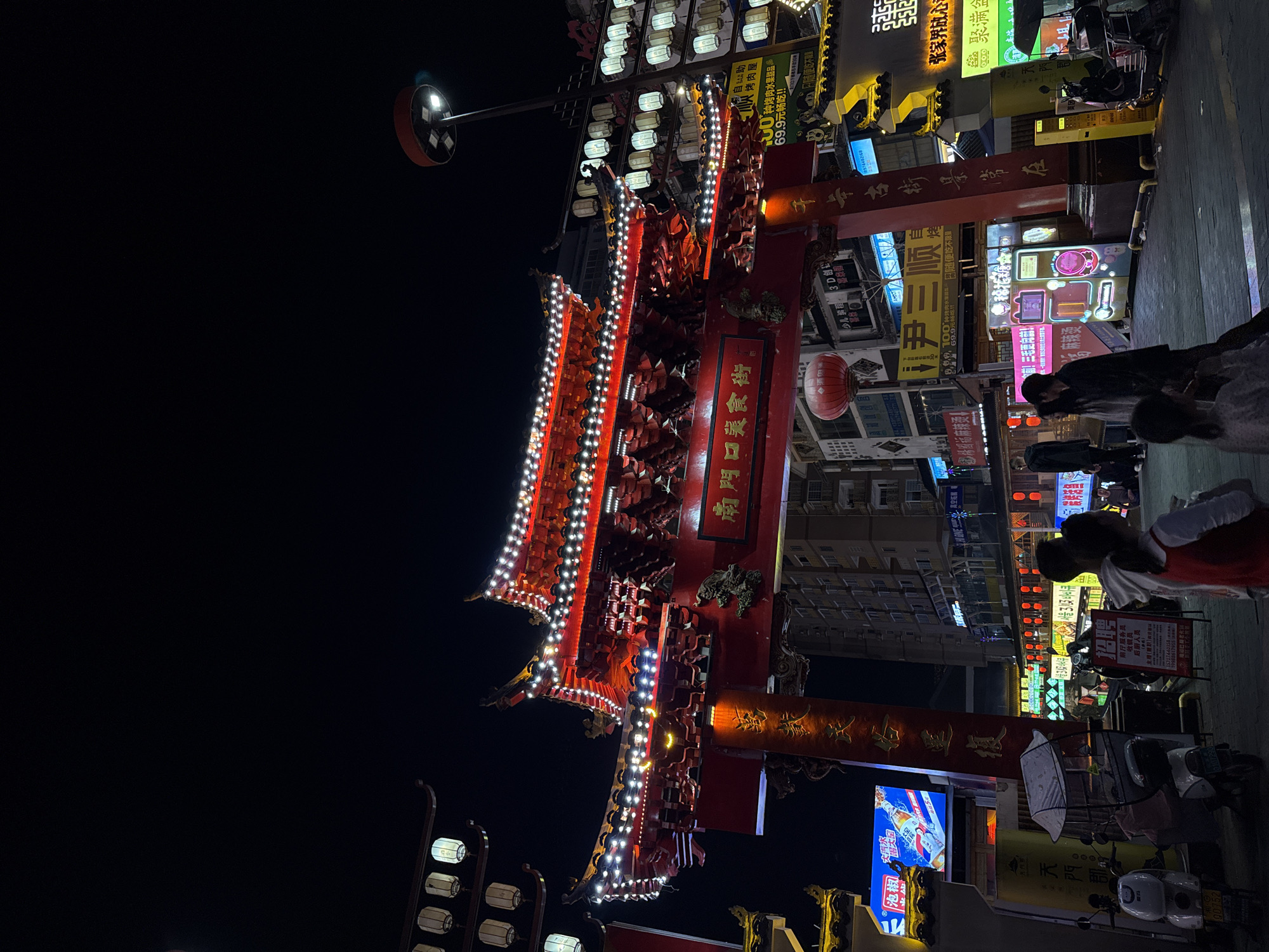 The image depicts a vibrant night scene on a bustling street in Zhangjiajie, China, centered around a brightly illuminated traditional Chinese archway (paifang). The sky is dark, indicating late evening or night, with numerous artificial lights illuminating the scene.
The dominant feature is a large, ornate red archway, intricately designed with traditional Chinese architectural elements and outlined with strings of white lights. On its central plaque, the text "南門口美食街" (Nanmenkou Food Street) is prominently displayed, suggesting the area is known for its culinary offerings. Chinese lanterns are visible hanging from the archway and nearby structures.
To the right of the archway, several people are seen walking or standing on the street, likely enjoying the evening atmosphere. Their attire is casual, suitable for an urban setting. A few motorcycles or scooters are parked along the side, common for local transportation.
The street is lined with multi-story buildings, their facades covered in a dense array of glowing commercial signs in various colors and styles. These signs advertise a wide range of businesses and services. Visible text from the signs includes "招聘" (Recruiting) for roles like "前厅服务员" (Front hall service staff), "收银员" (Cashier), and "后厨人员" (Back kitchen staff). Specific establishments mentioned are "楊阁福麻辣烫" (Yanggefu Malatang) and "自助烤肉屋" (Self-service BBQ house), with an offer for "20种烤肉水果甜品 69.9元畅吃!!" (20 kinds of BBQ, fruits, desserts, eat all you can for 69.9 yuan!!). Other texts like "张家界放心" (Zhangjiajie assured) and "少儿编程 3D创" (Children's programming 3D creation) are also present, providing diverse commercial context. A street light with multiple bright bulbs extends from the upper left.
The overall impression is one of a lively, energetic commercial district, likely popular for dining and entertainment, illuminated against the night sky.