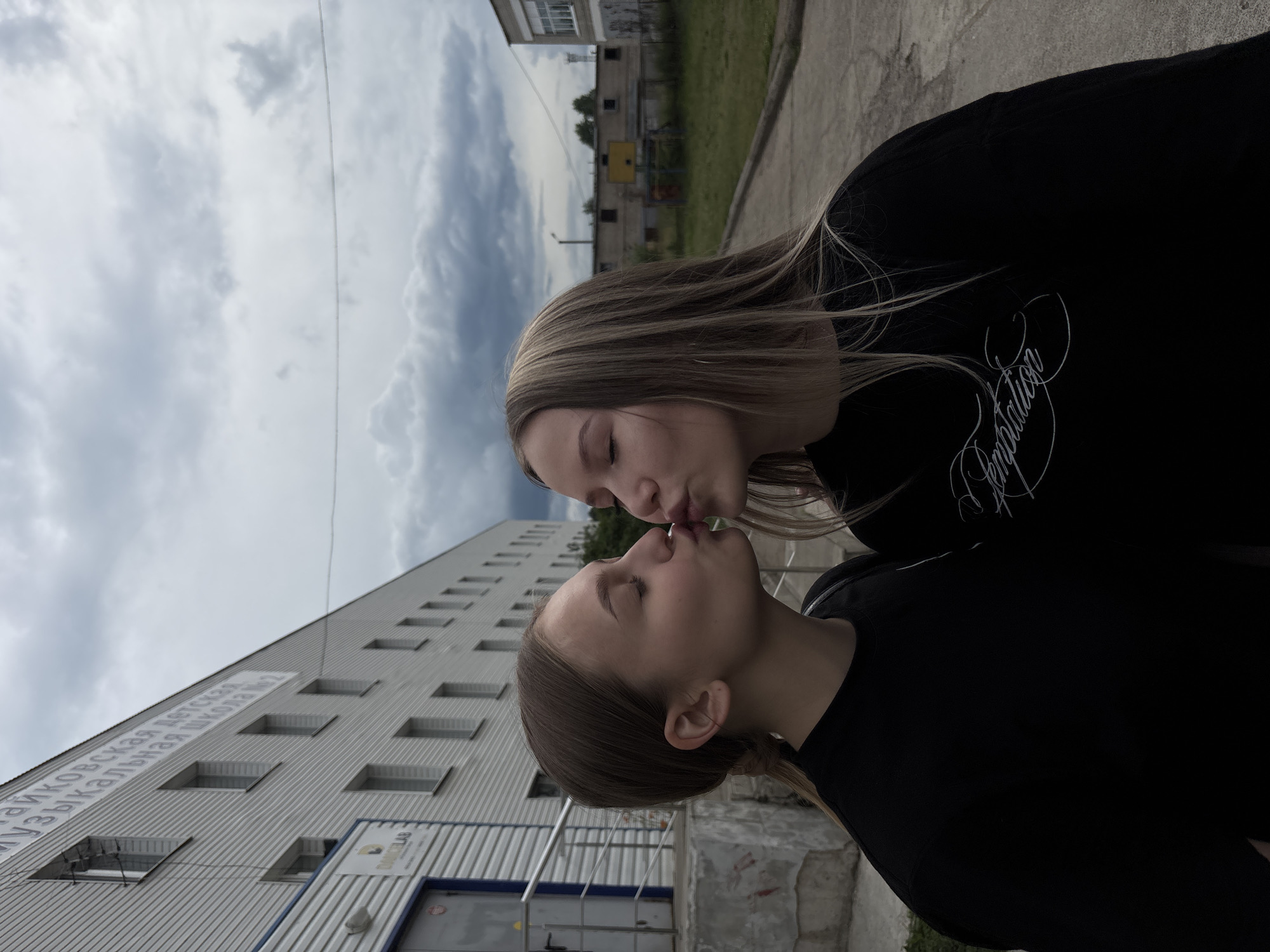 This is a vertical, outdoor, full-shot photograph of two young women, seemingly teenagers, about to kiss. Both are wearing black tops and have light brown hair. The woman in the foreground is closer to the camera, and her eyes are closed. The woman behind her, who is partially obscured, has her eyes closed as well, leaning in for a kiss.
The background features a long, light-colored building with multiple windows and horizontal siding. A large banner with Russian text is visible on the side of the building. The sky above is overcast with dramatic clouds, suggesting either early morning or late afternoon, possibly with impending rain. The ground appears to be asphalt or concrete. A yellow structure, possibly a playground element, is visible in the distance behind the building. The overall setting suggests an urban or suburban environment in Chaikovsky, Russia. The emotion conveyed is romantic or affectionate.
The visible text on the banner on the building is in Russian and appears to read "ЧАЙКОВСКАЯ ДЕТСКАЯ МУЗЫКАЛЬНАЯ ШКОЛА № 2," which translates to "Chaikovsky Children's Music School No. 2." There is also some text on a smaller sign on the building that is difficult to read. The woman behind has text on her shirt that reads "Temptation" in a cursive font.