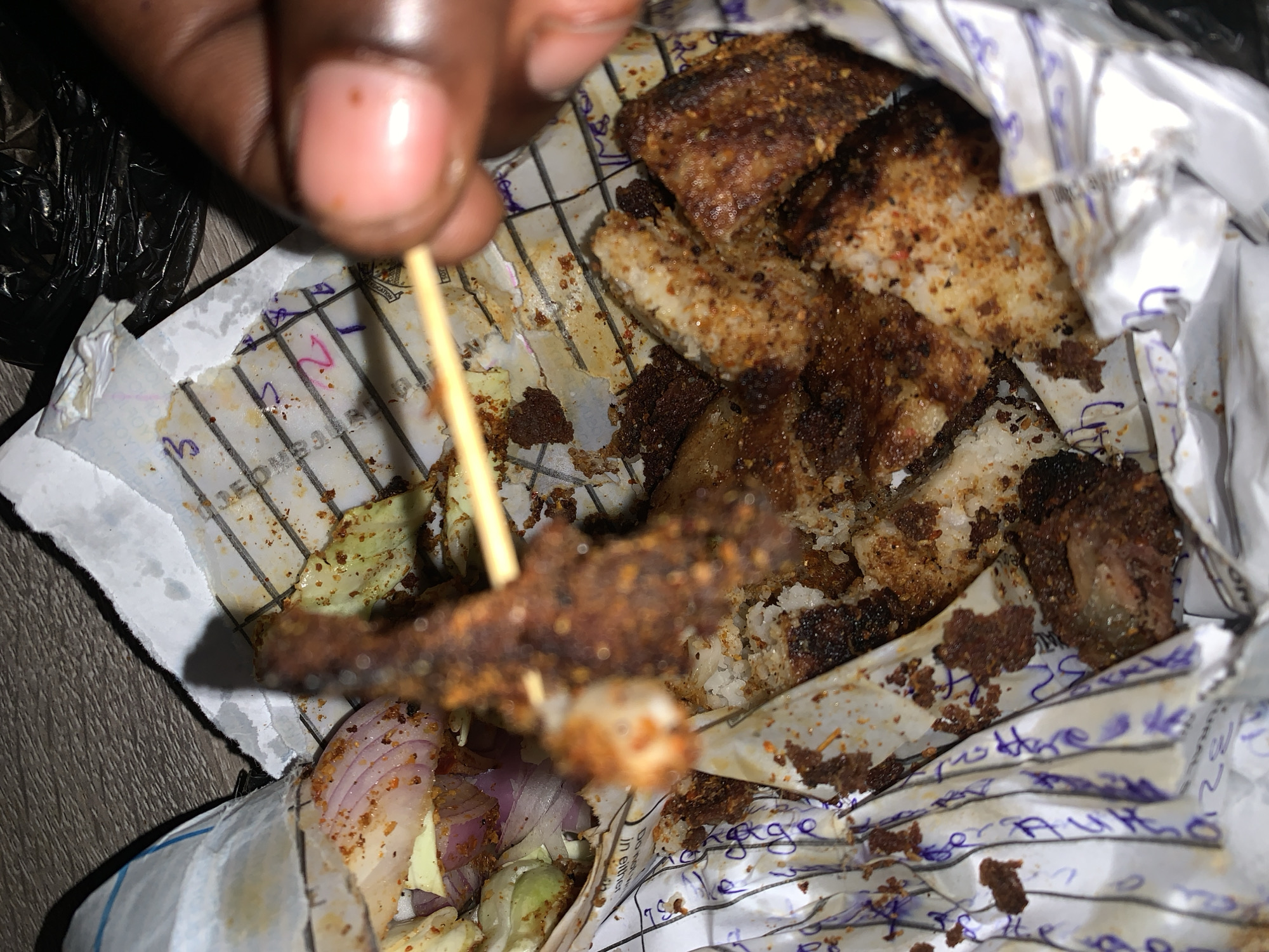 A close-up, overhead shot shows a person's hand holding a skewer with fried food. The food appears to be chicken or fish coated in spices and is served in a paper wrapper. There are also pieces of red onion visible. The food is partially obscured by the wrapper and a black plastic bag. The background is a wooden surface. The scene is indoors, possibly a restaurant or street food vendor in New Stadium, Nigeria.