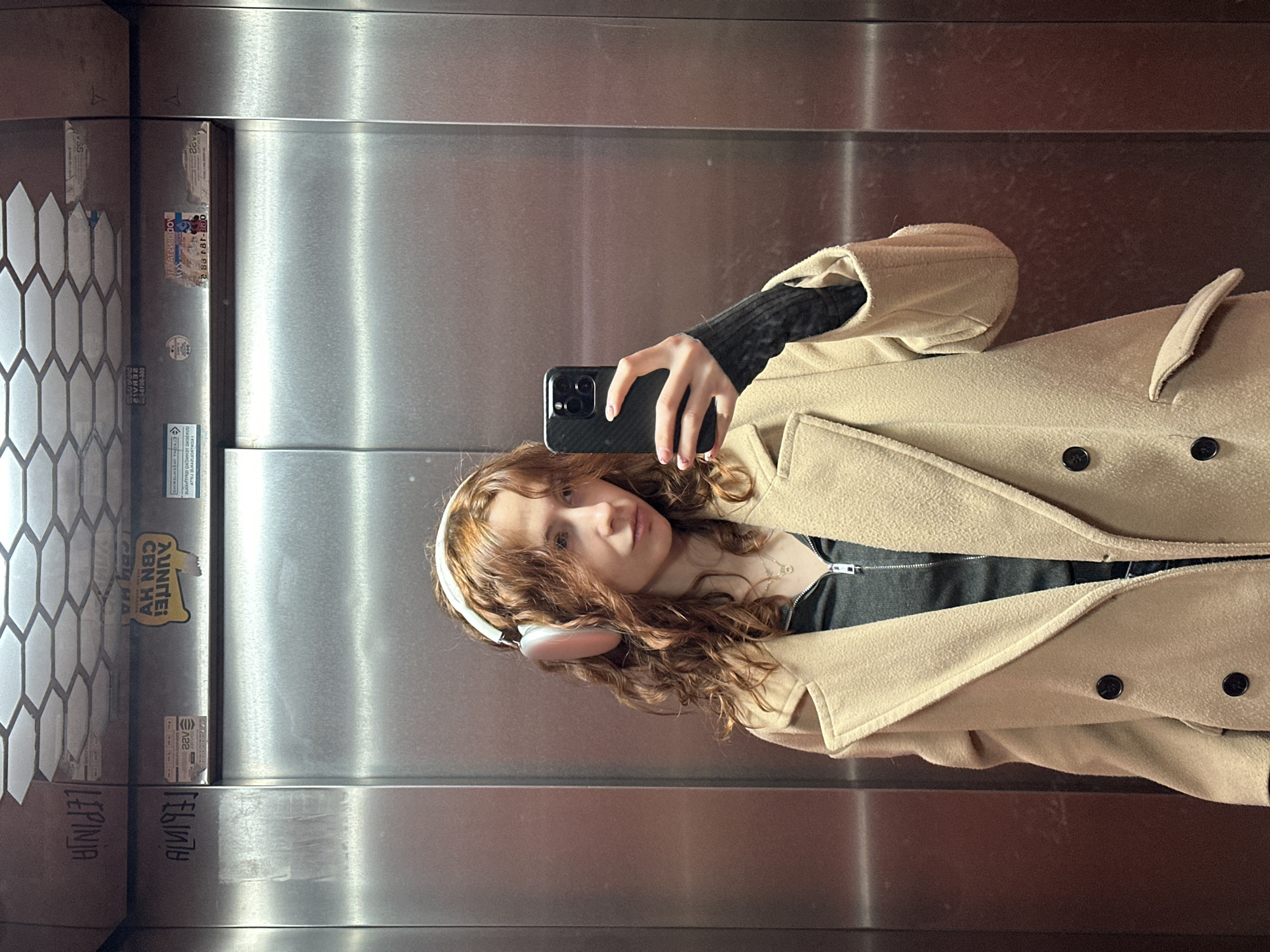 A woman is taking a selfie in an elevator. She is wearing a beige coat, a black top, and white headphones. Her hair is curly and red. She is holding a black smartphone. The elevator has stainless steel walls. On the left side of the elevator, there are some stickers and writings. The location is identified by "LEPINJA" written on the elevator wall, which is a known street food in Novi Sad, Serbia. Therefore, the location is presumed to be in Novi Sad, Serbia.